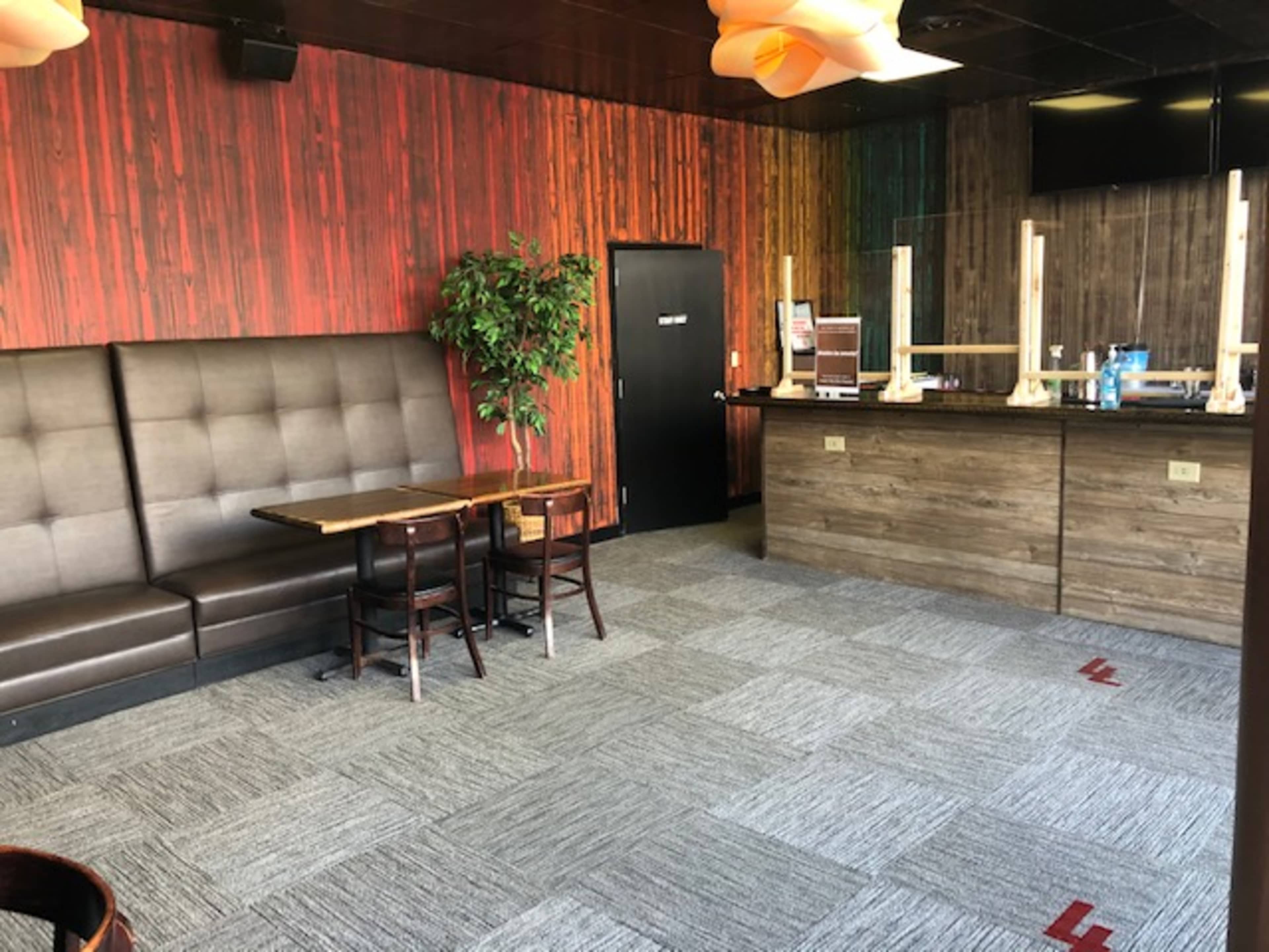 An interior of a restaurant or bar featuring a brown leather booth, wooden tables, a plant, and a dark wooden counter with a television on the wall.