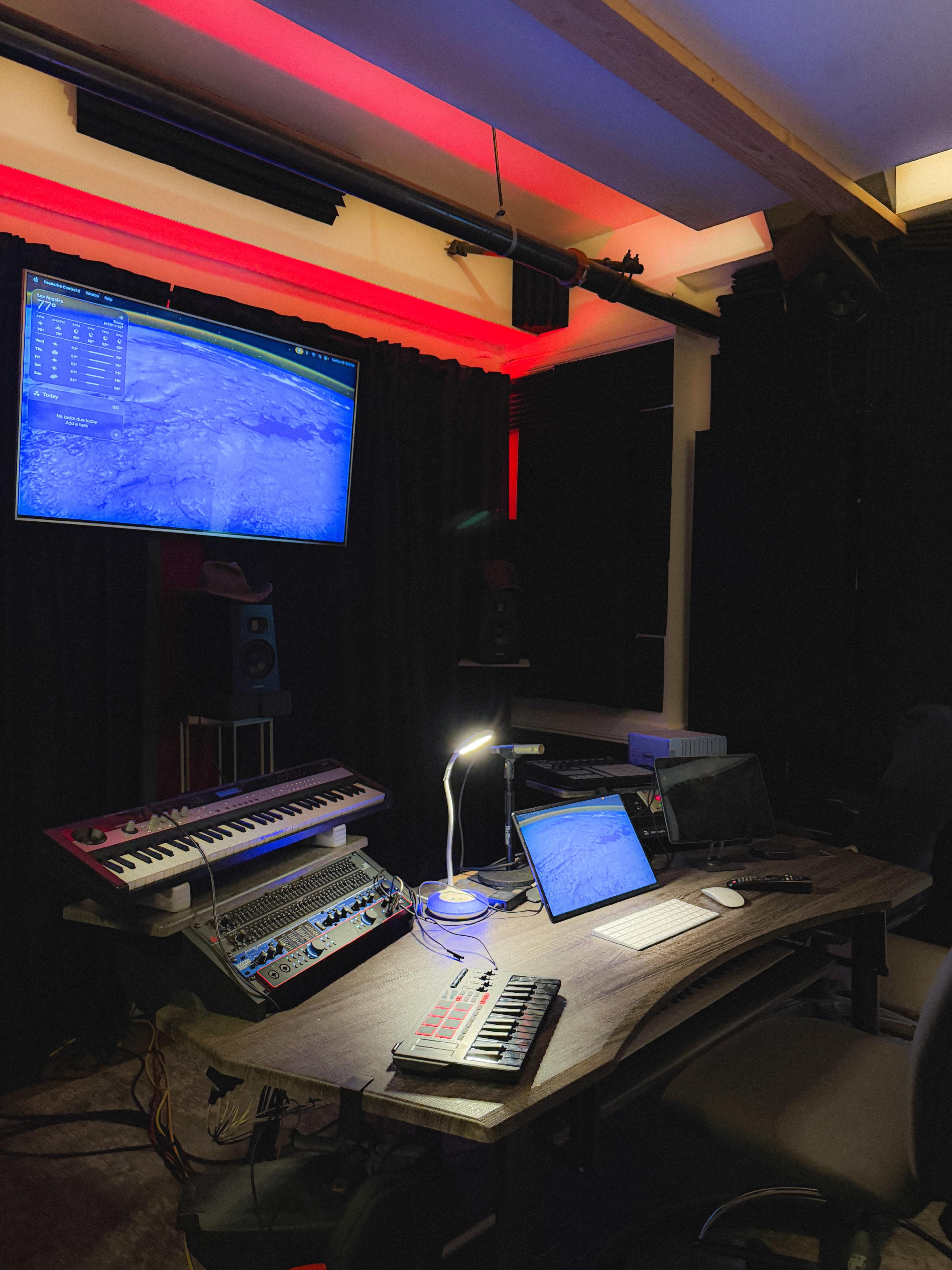 The image shows a music studio setup with a computer, keyboard, and various electronic instruments, all arranged on a wooden desk in a darkened room with red accent lighting.