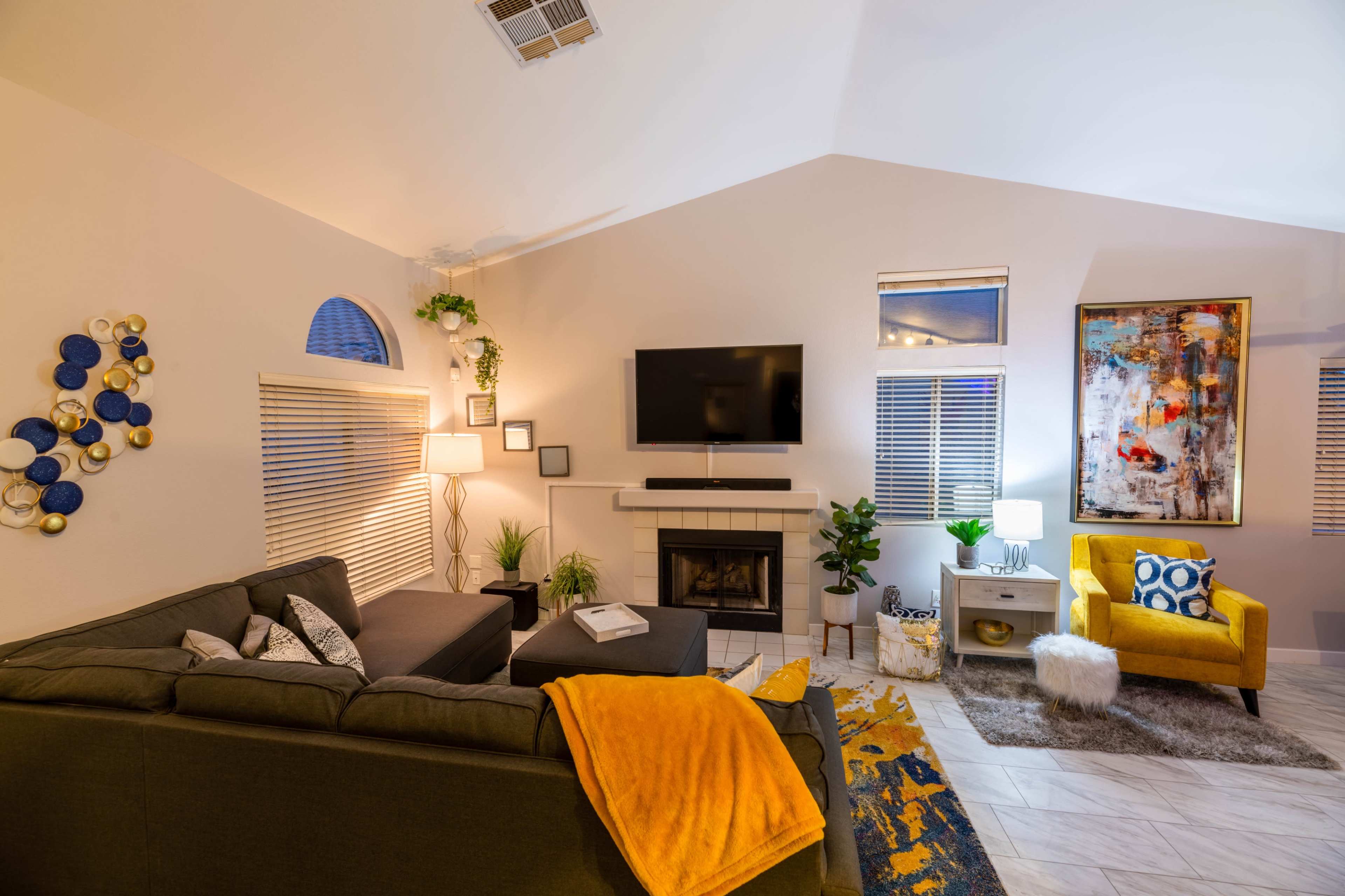 A modern living room features a gray sofa, yellow accent chair, fireplace, and a large wall-mounted television.