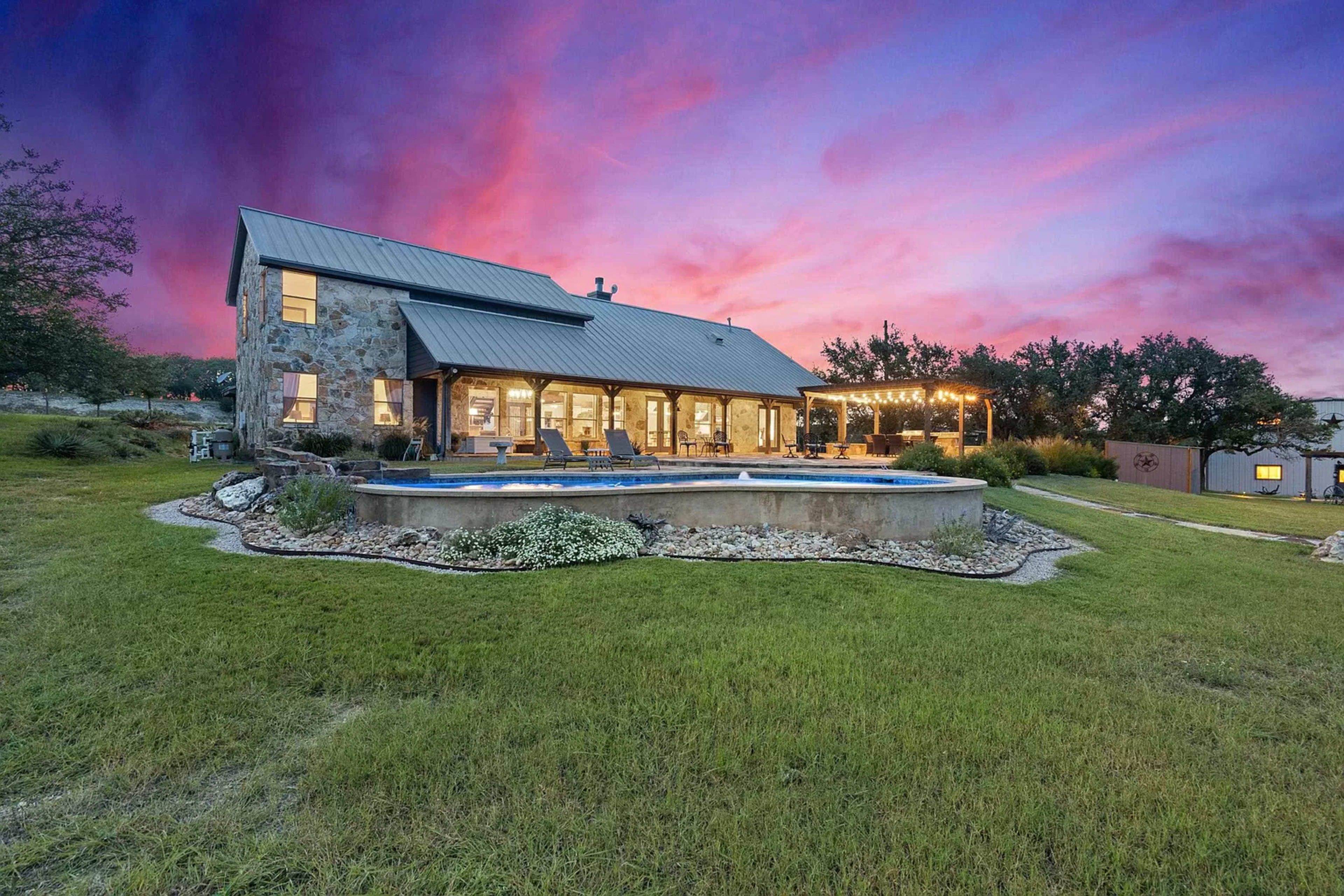 A stone house with a metal roof is situated near a pool, surrounded by green grass and evening skies painted in shades of pink and purple.