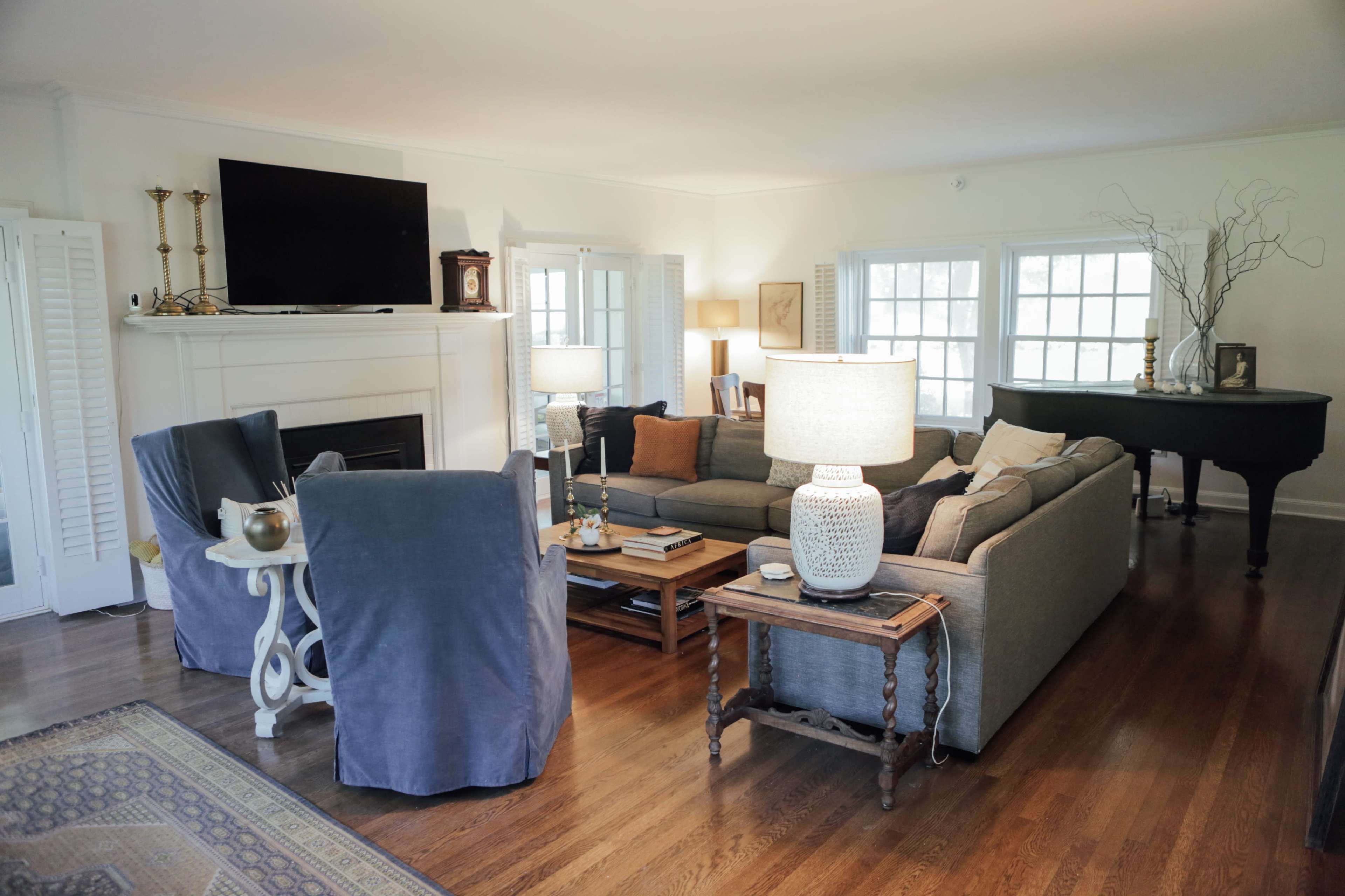 The living room features a gray sectional sofa, two blue chairs, a coffee table, and a grand piano, with natural light streaming through multiple windows.