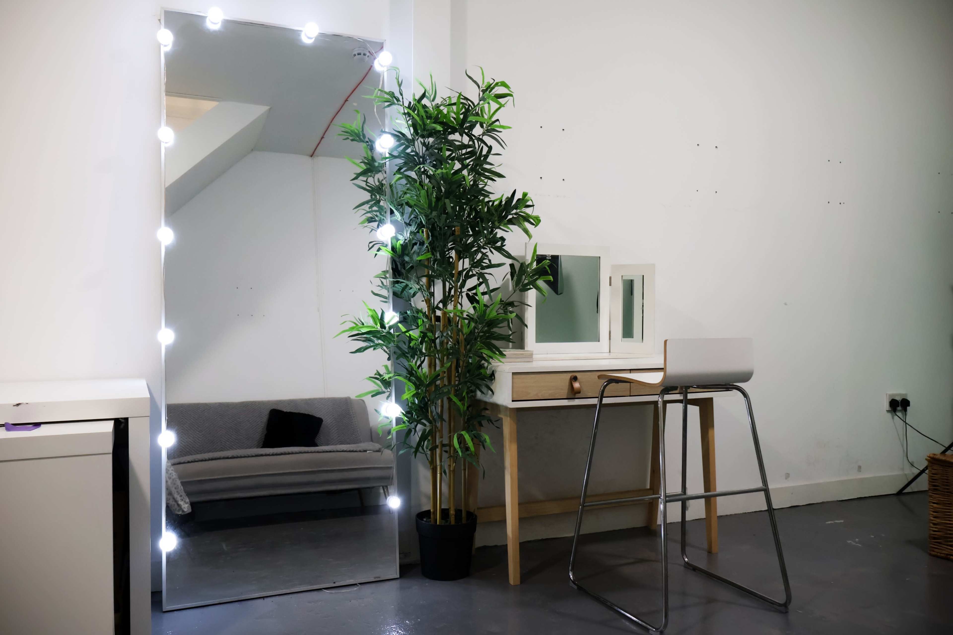 The image shows a well-lit dressing area featuring a full-length mirror with lights, a potted plant, and a light-colored vanity table with a stool.