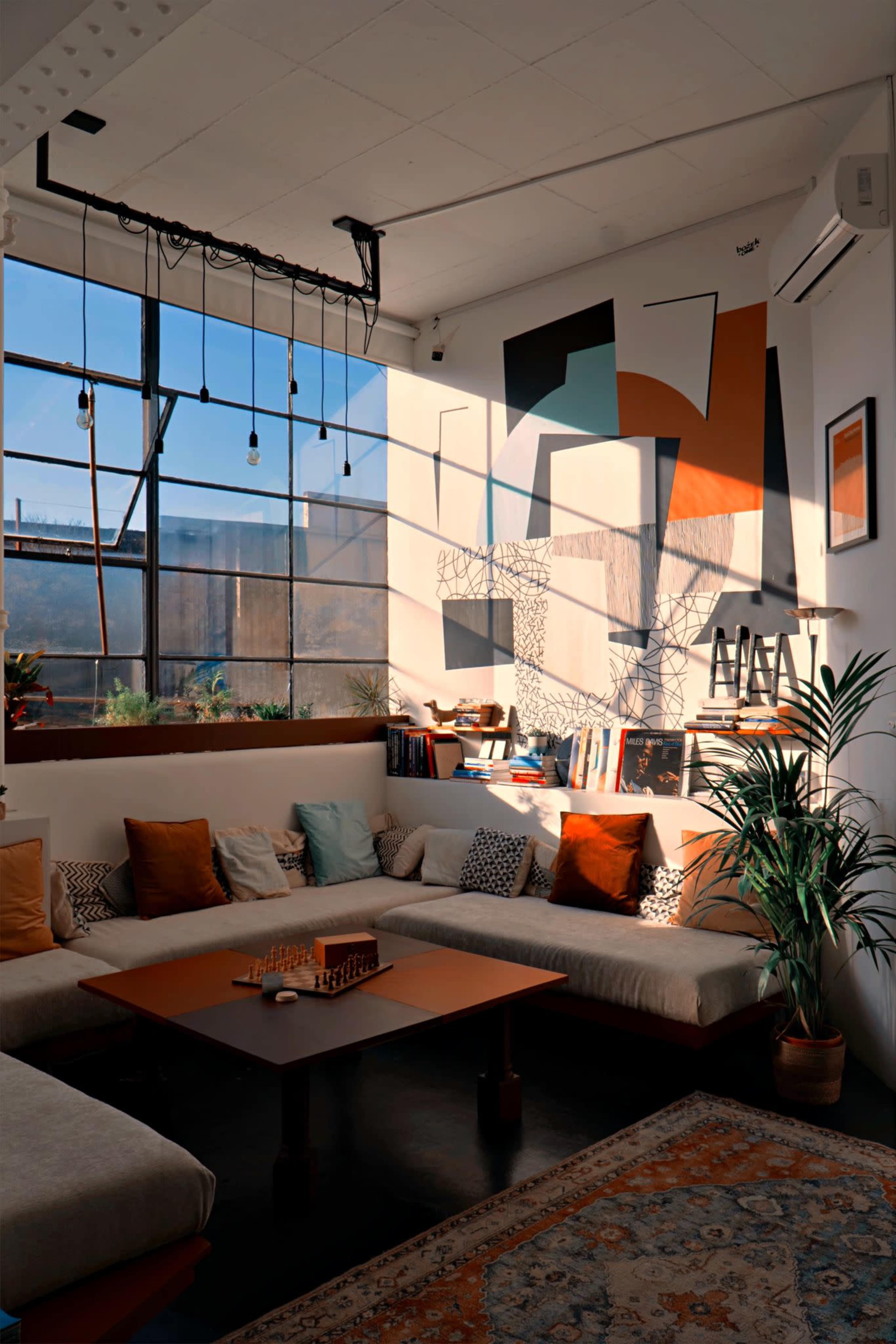 The image shows a cozy living room with a large window casting geometric shadows, a sectional sofa with various cushions, and a wooden coffee table with a chessboard on it.