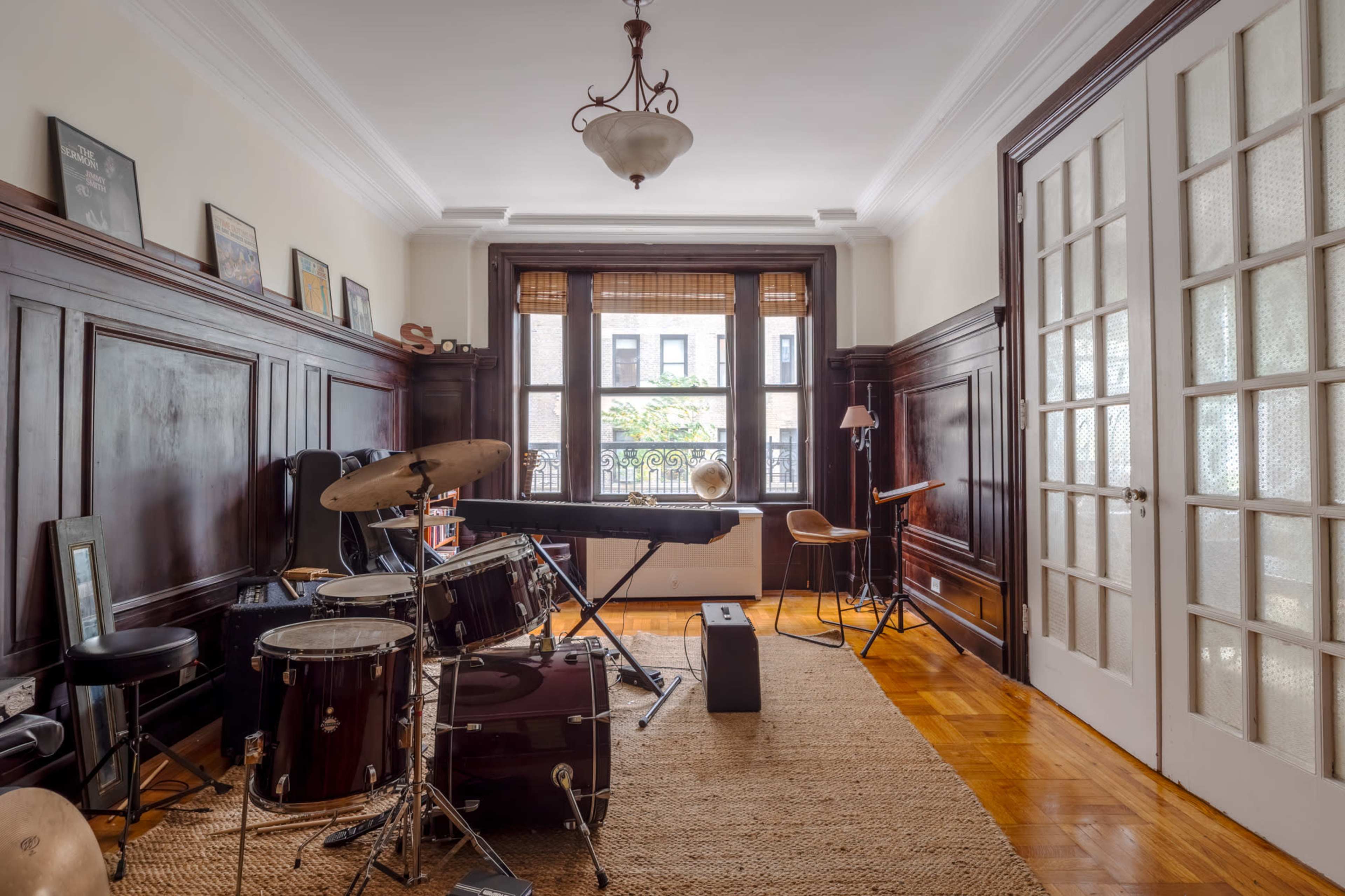 The image shows a music room featuring a drum set, keyboard, and various instruments against a backdrop of dark wooden paneling and large windows.