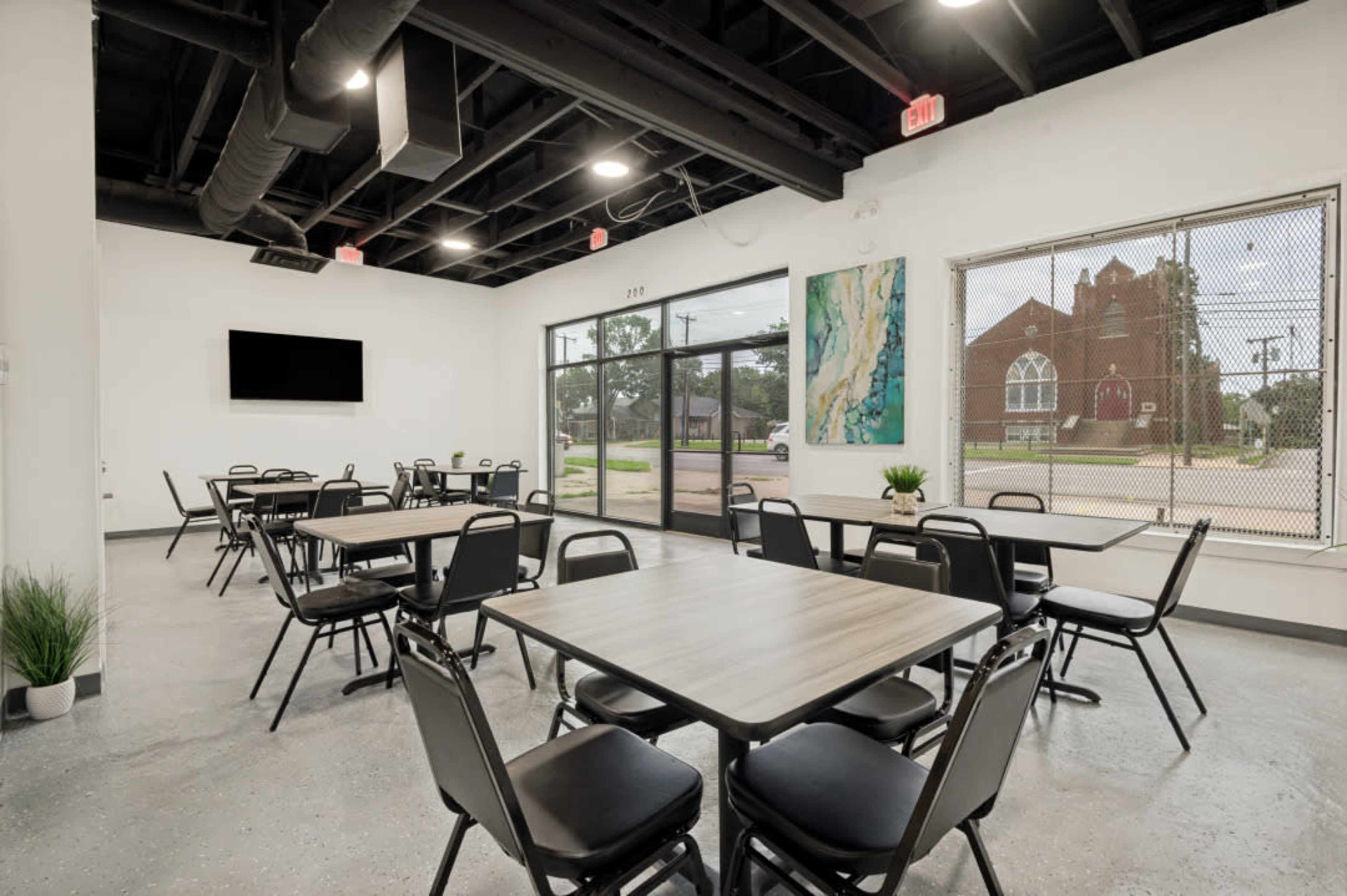 The image shows a modern dining or meeting space featuring several rectangular tables and chairs, with large windows allowing natural light and a view of an adjacent building.