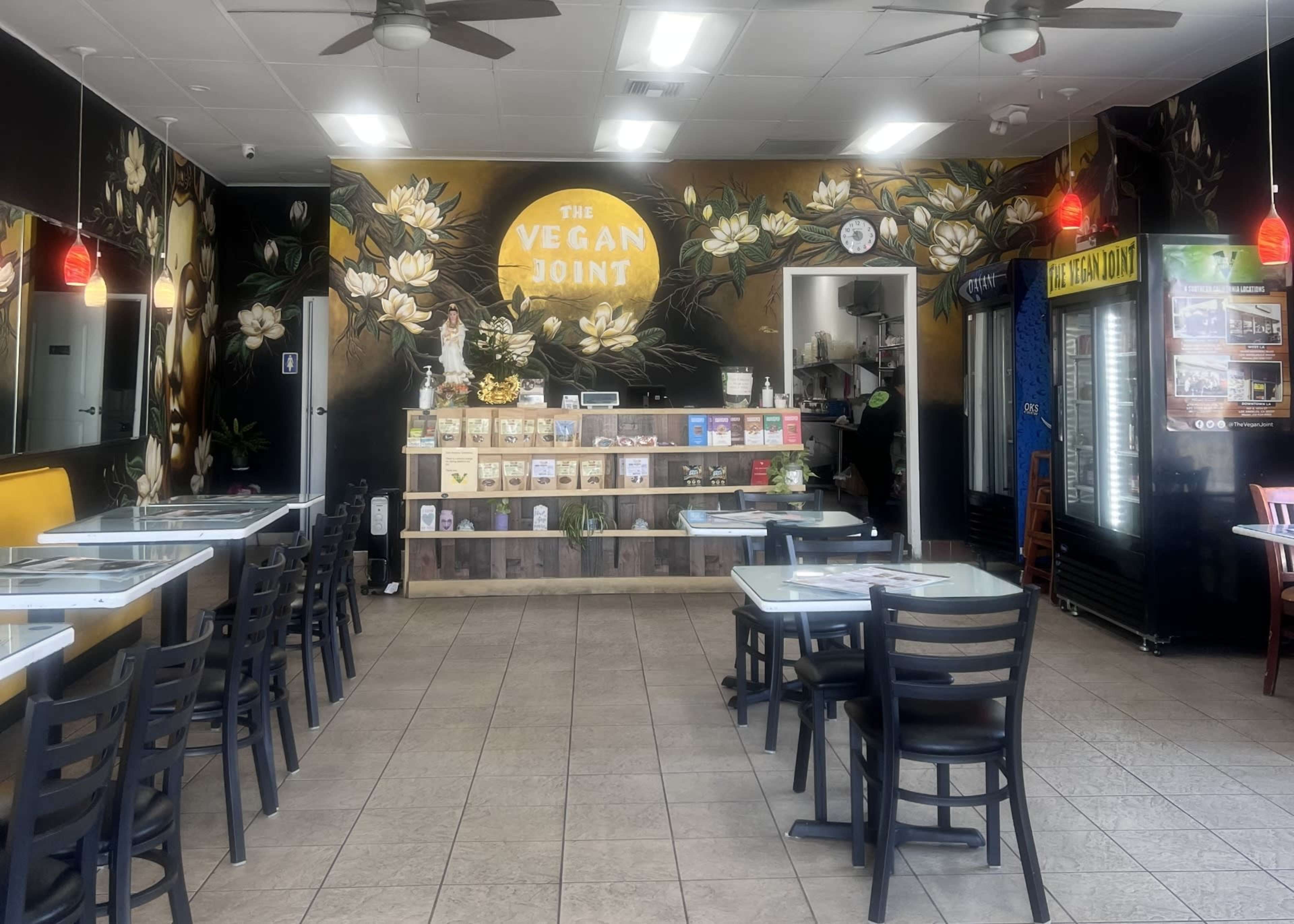 The interior of a vegan restaurant features a brightly colored mural, wooden display shelf filled with products, and seating arranged around tables.