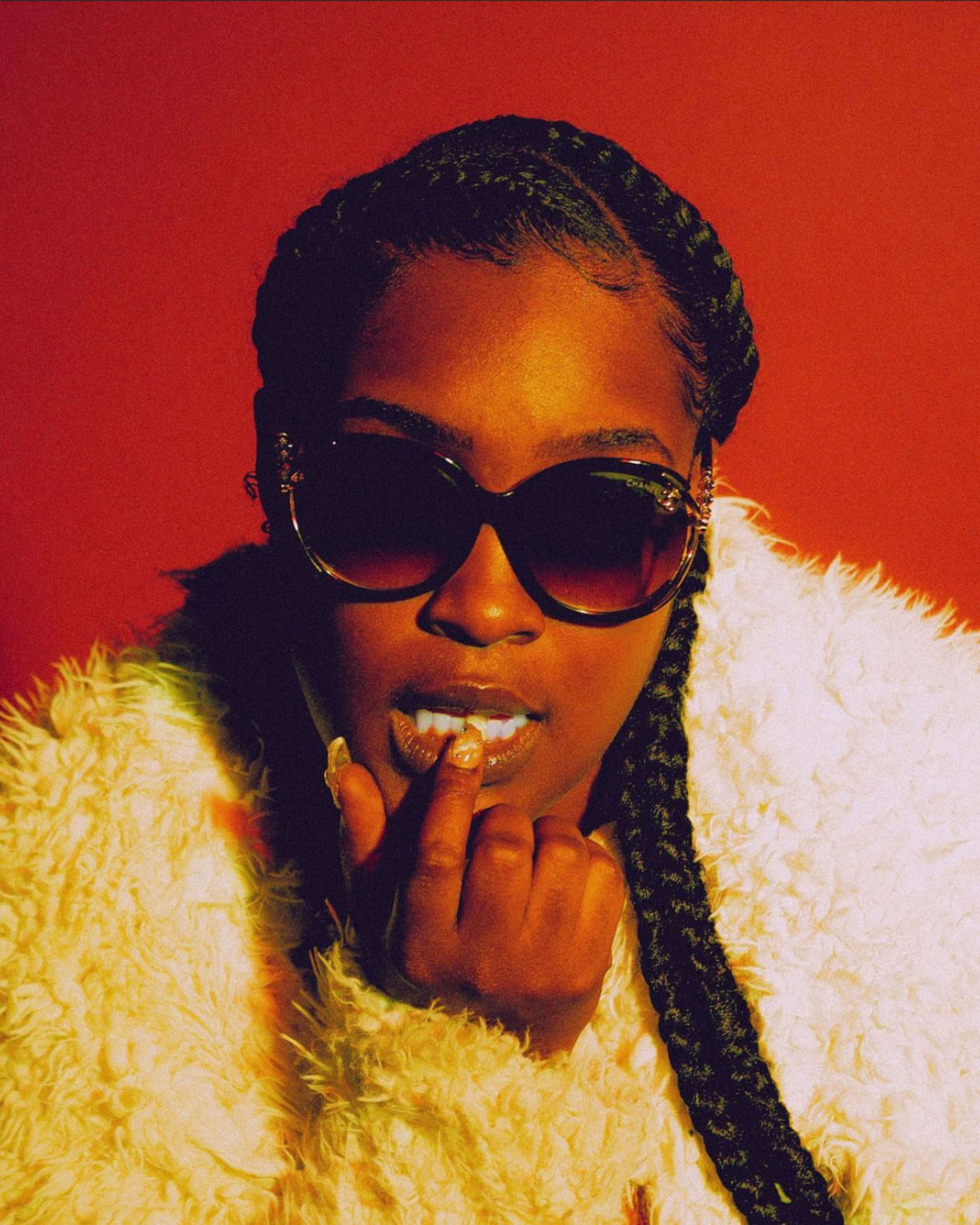 A person with braided hair is wearing large sunglasses and a fluffy white coat, posing against a red background while gesturing toward their lips.