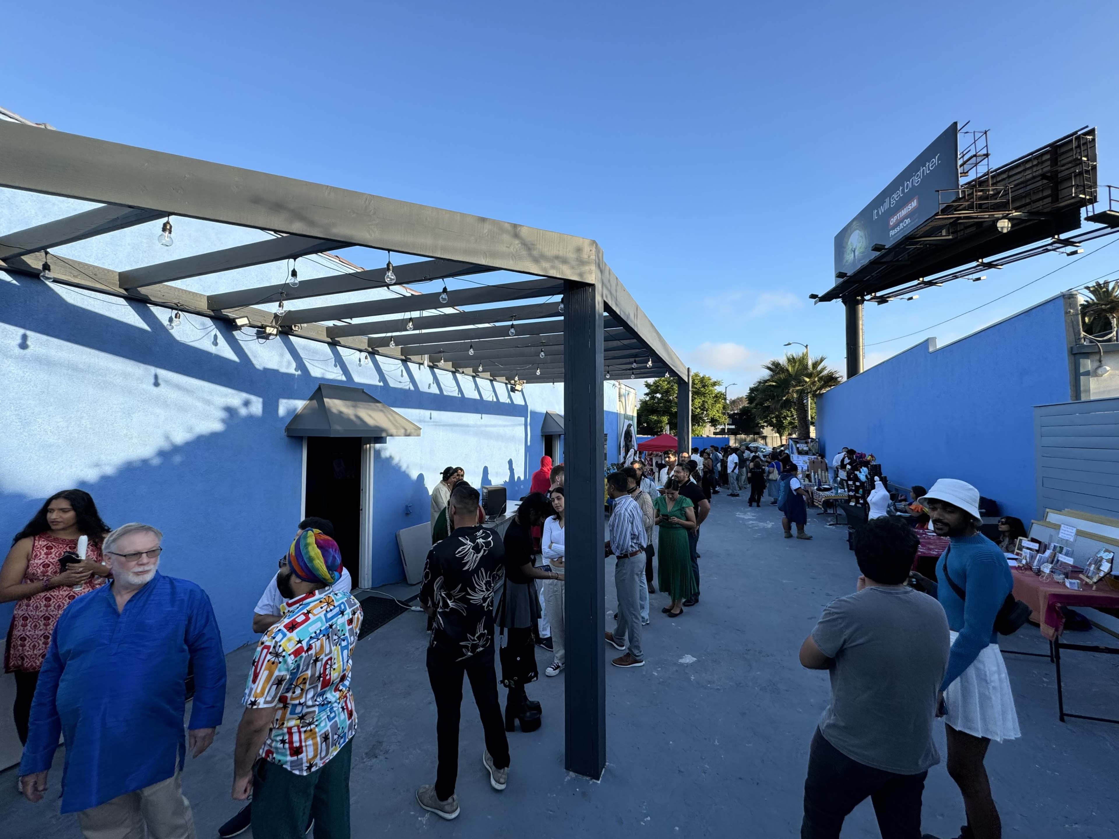 A crowd of people gathers in a sunny outdoor space beside a blue building, with some waiting in line and others engaging in conversation.