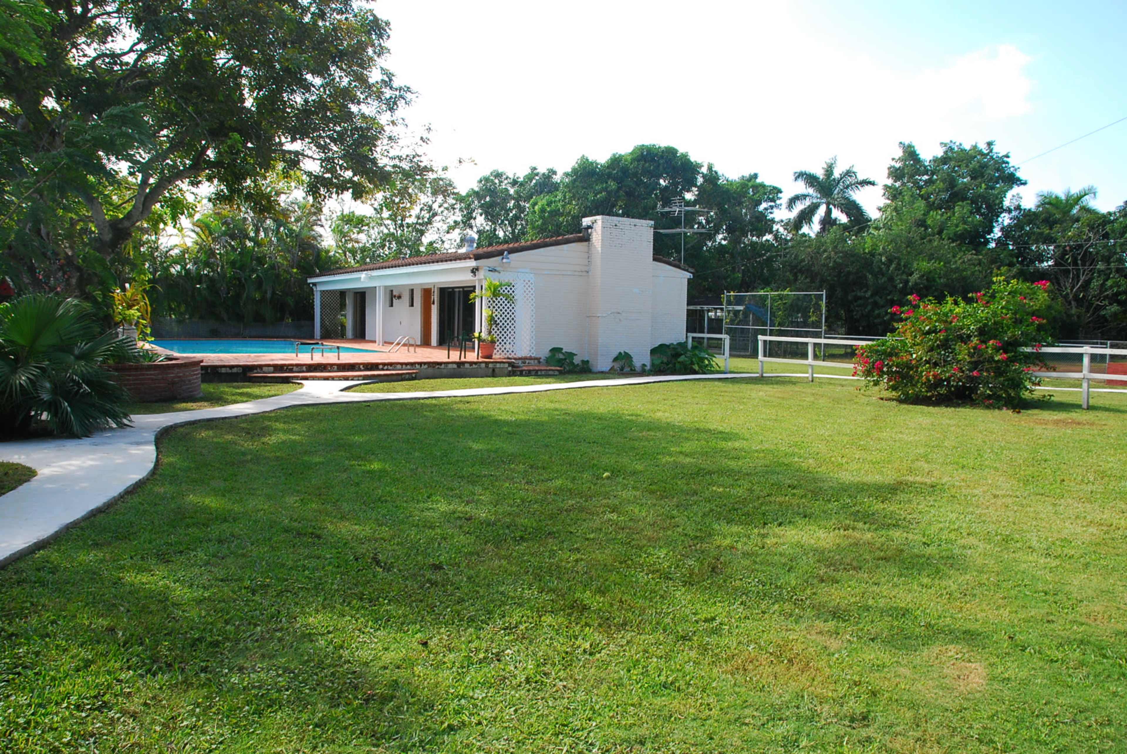 The image shows a spacious backyard with neatly manicured grass, a house partially visible in the background, a swimming pool, and surrounding greenery.
