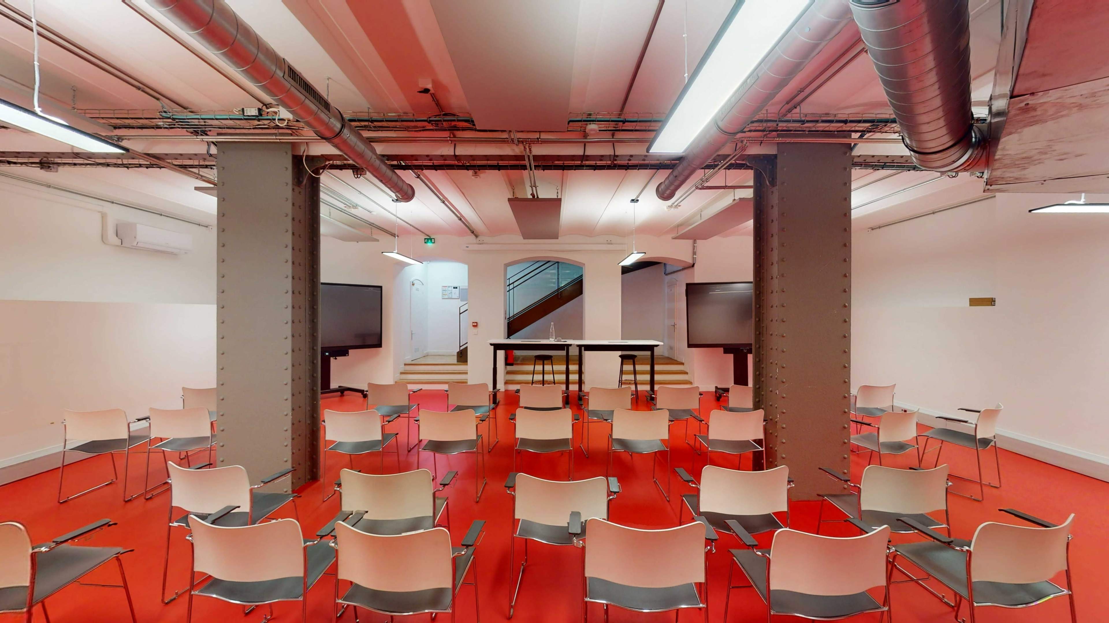 An empty room with red flooring, featuring rows of white chairs arranged for an event, with a staircase and two large screens at the front.