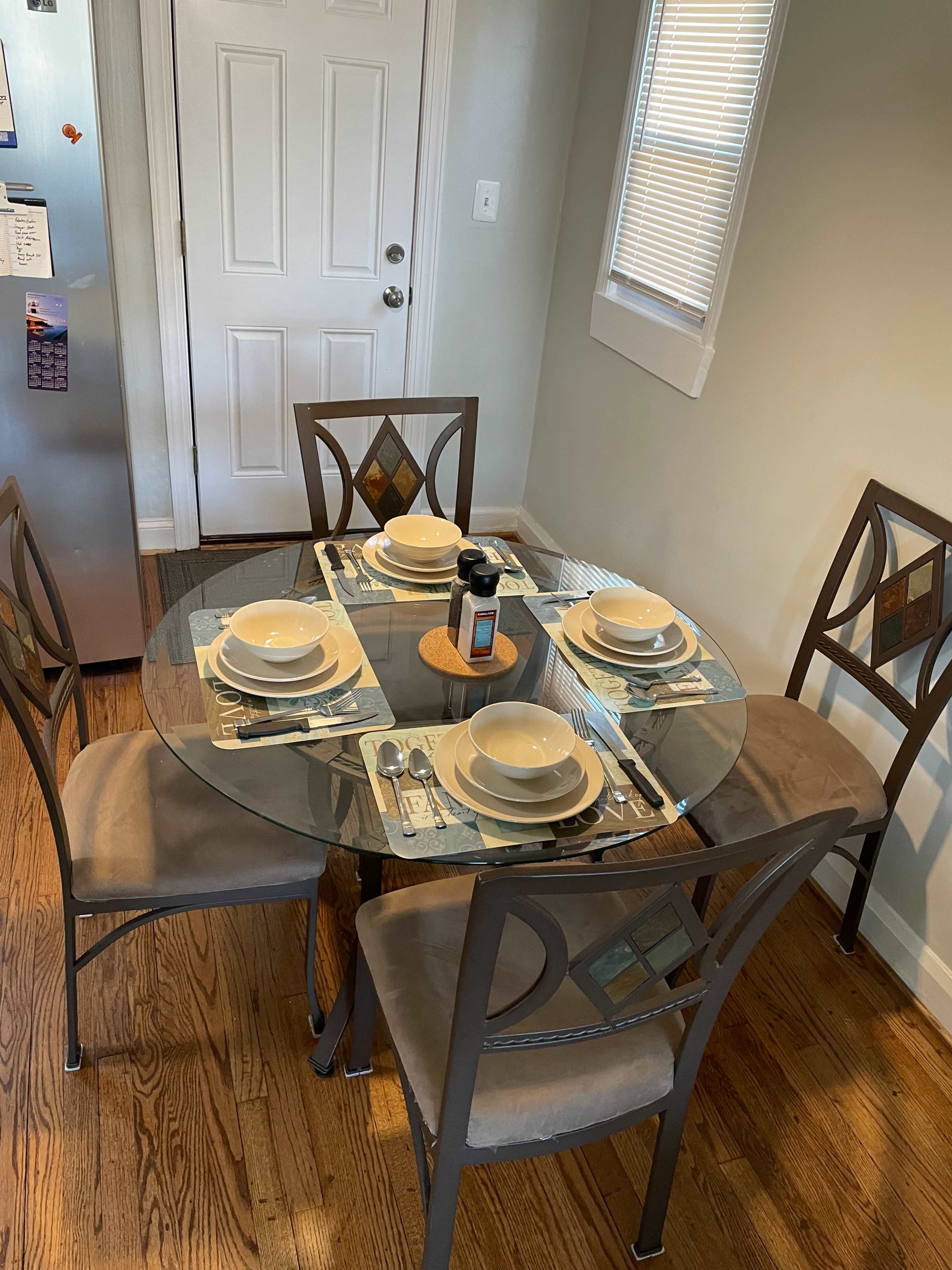 A round glass dining table is set with four bowls, utensils, and a small condiments container, surrounded by four chairs in a well-lit kitchen.