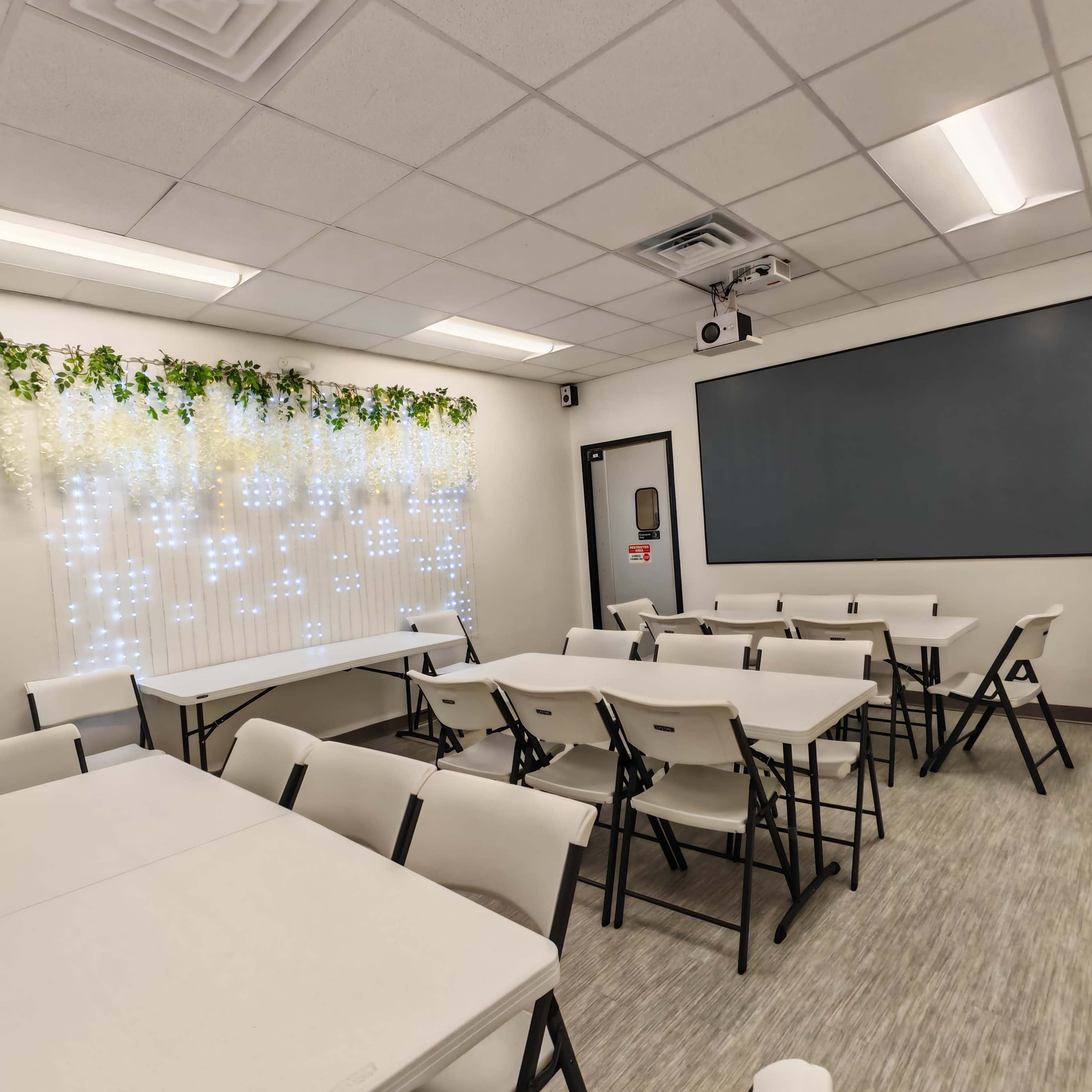 The image shows a classroom with several folding tables and chairs arranged in rows, along with decorative greenery and fairy lights along one wall.