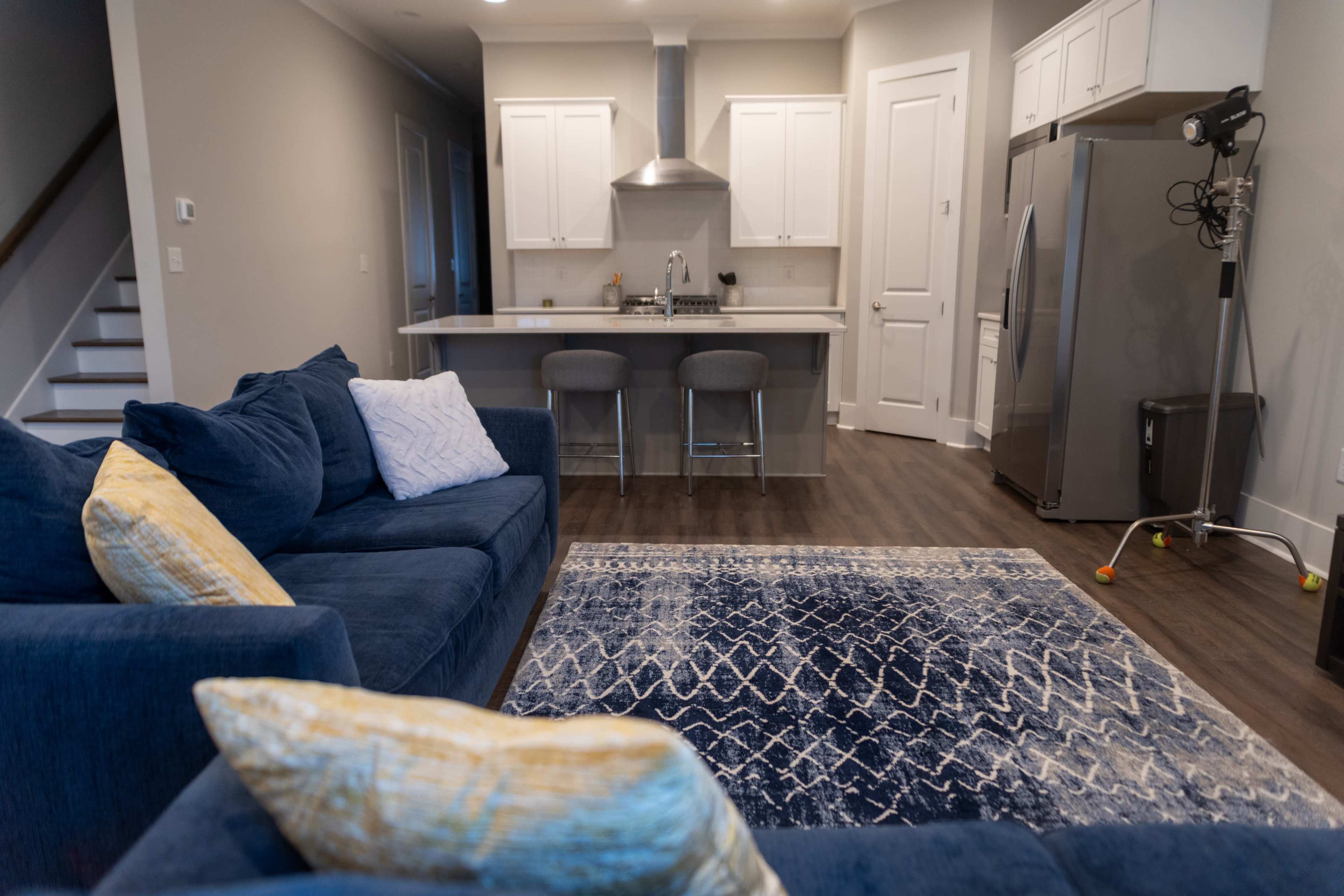 The image shows a modern living room with a blue sectional sofa, a decorative rug, and a kitchen area featuring stainless steel appliances and a countertop.