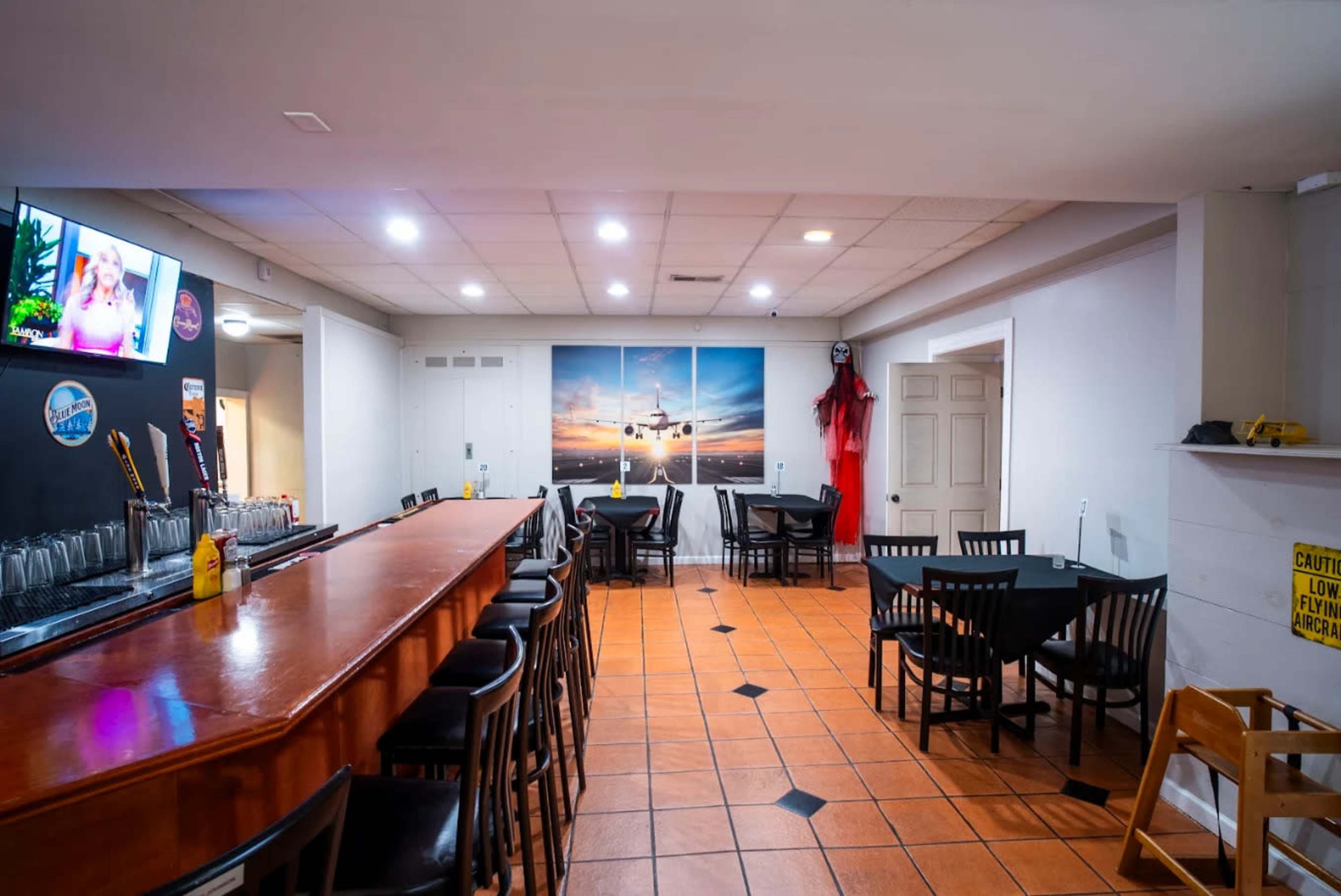The image shows a spacious restaurant interior with a wooden bar, black tables and chairs, and artwork featuring a sunset on the wall.