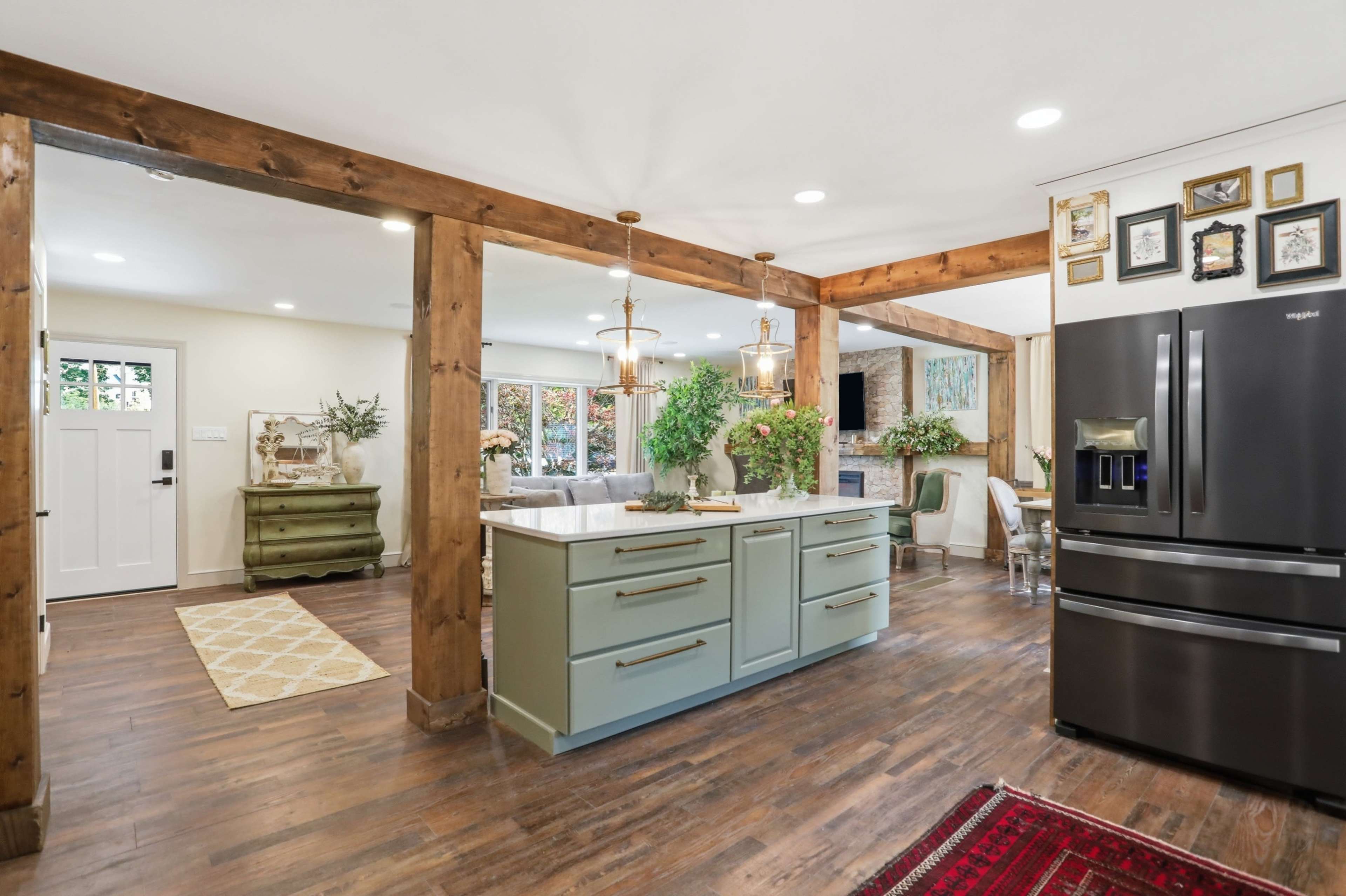The image shows a modern kitchen with an island in soft green, wooden beams, and a black refrigerator, connecting to a cozy dining area with large windows.