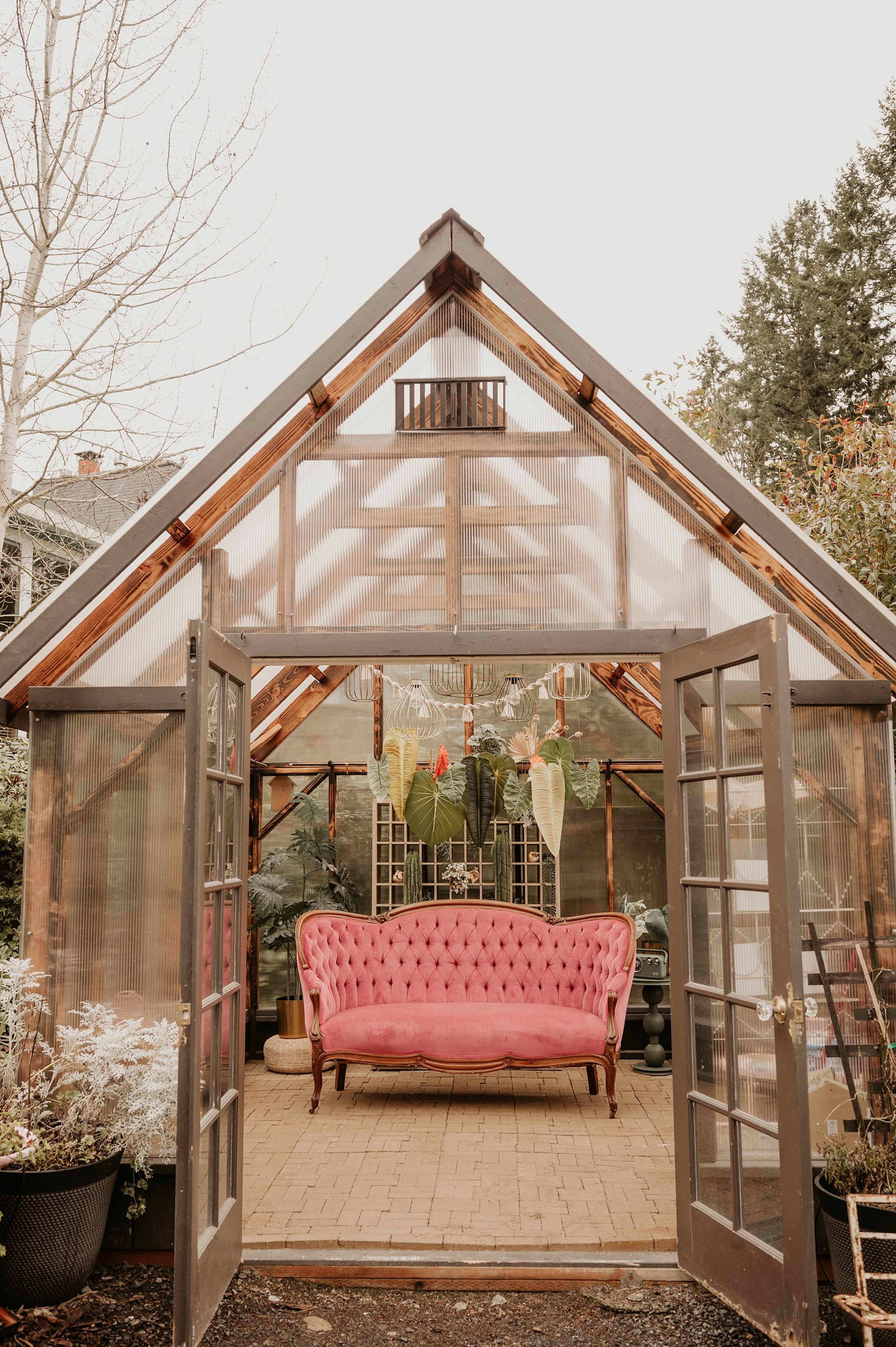 A pink tufted sofa sits inside a greenhouse, surrounded by various plants and large glass doors leading outside.