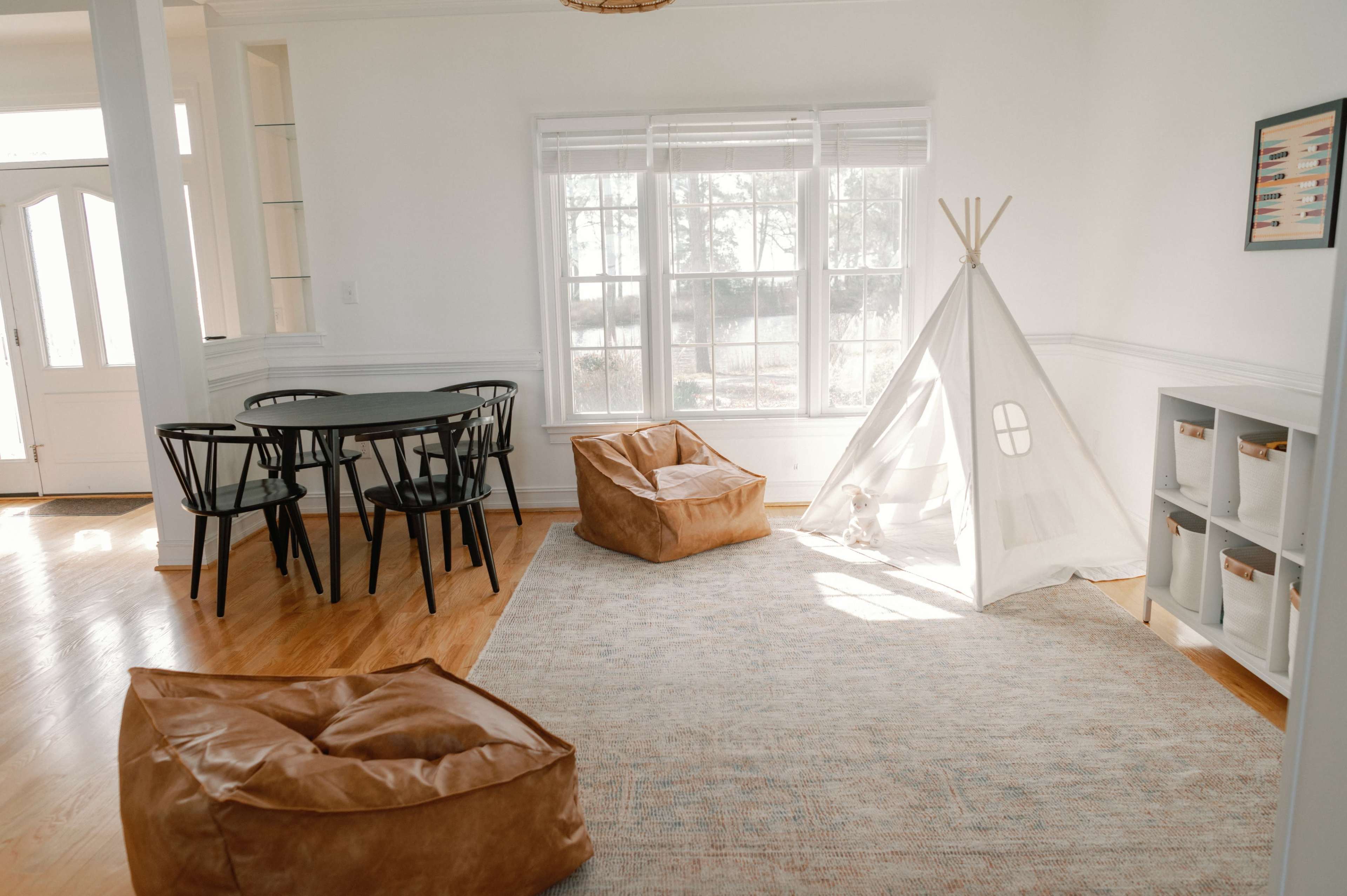 A bright room features a table with chairs, two bean bag chairs, a light-colored rug, and a children's teepee by a large window.