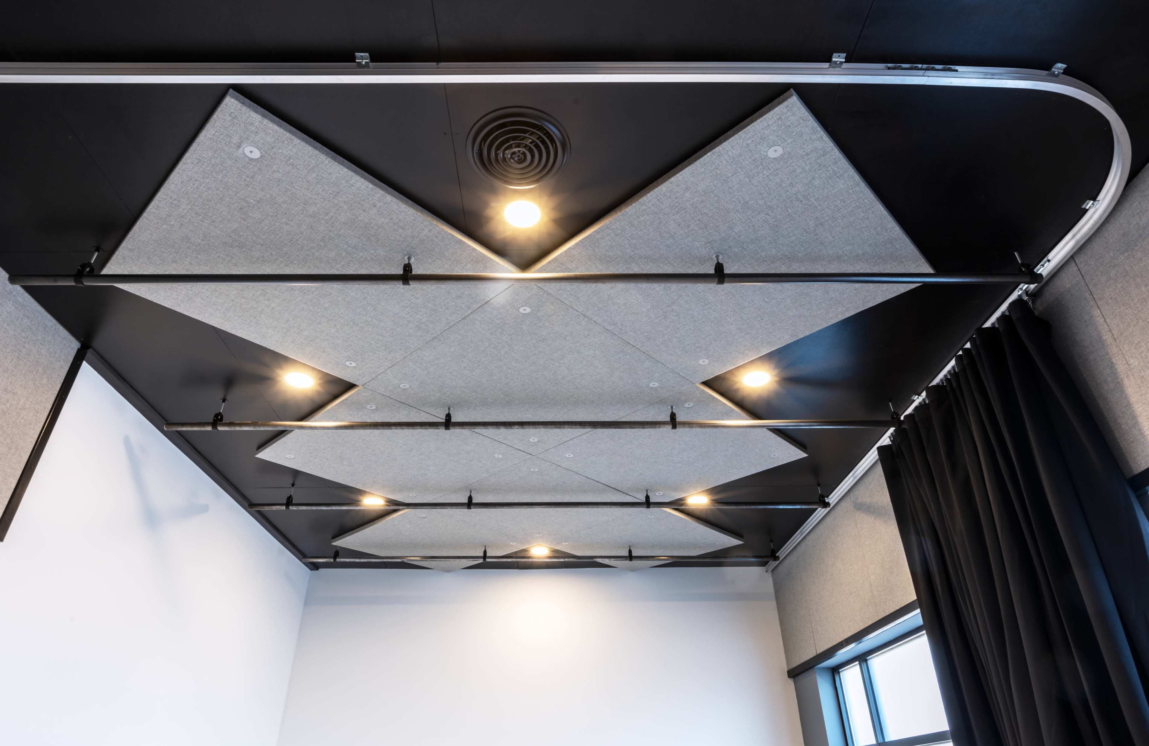 A ceiling with triangular acoustic panels arranged in a pattern, accompanied by recessed lights and a black curtain on one side.