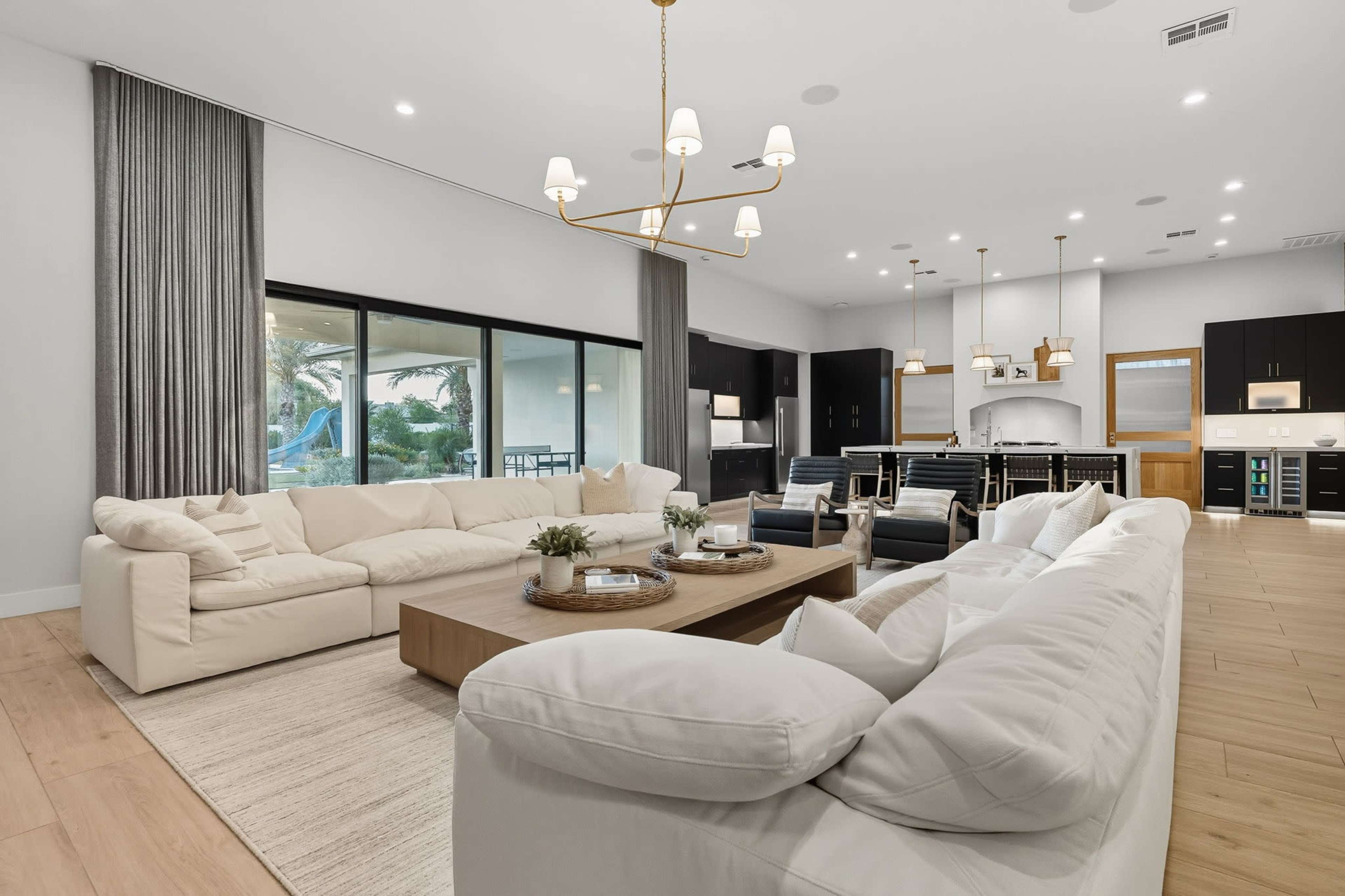 The image shows a spacious living area with a large sectional sofa, a wooden coffee table, and a dining area connected to a modern kitchen, all featuring neutral colors and abundant natural light.