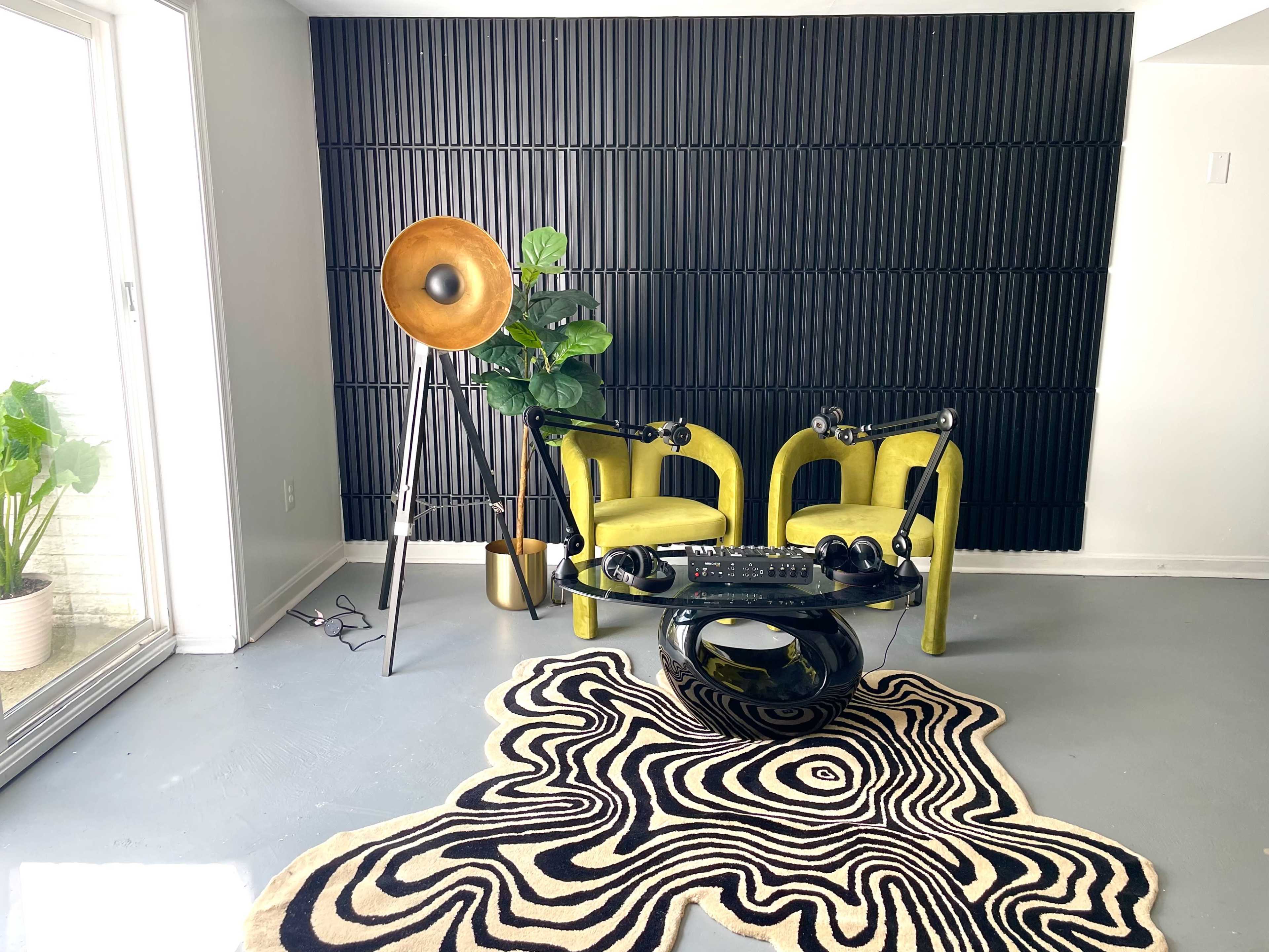 A contemporary living space featuring two green chairs, a black and white patterned rug, a tall floor lamp, and a potted plant, all against a textured black wall.