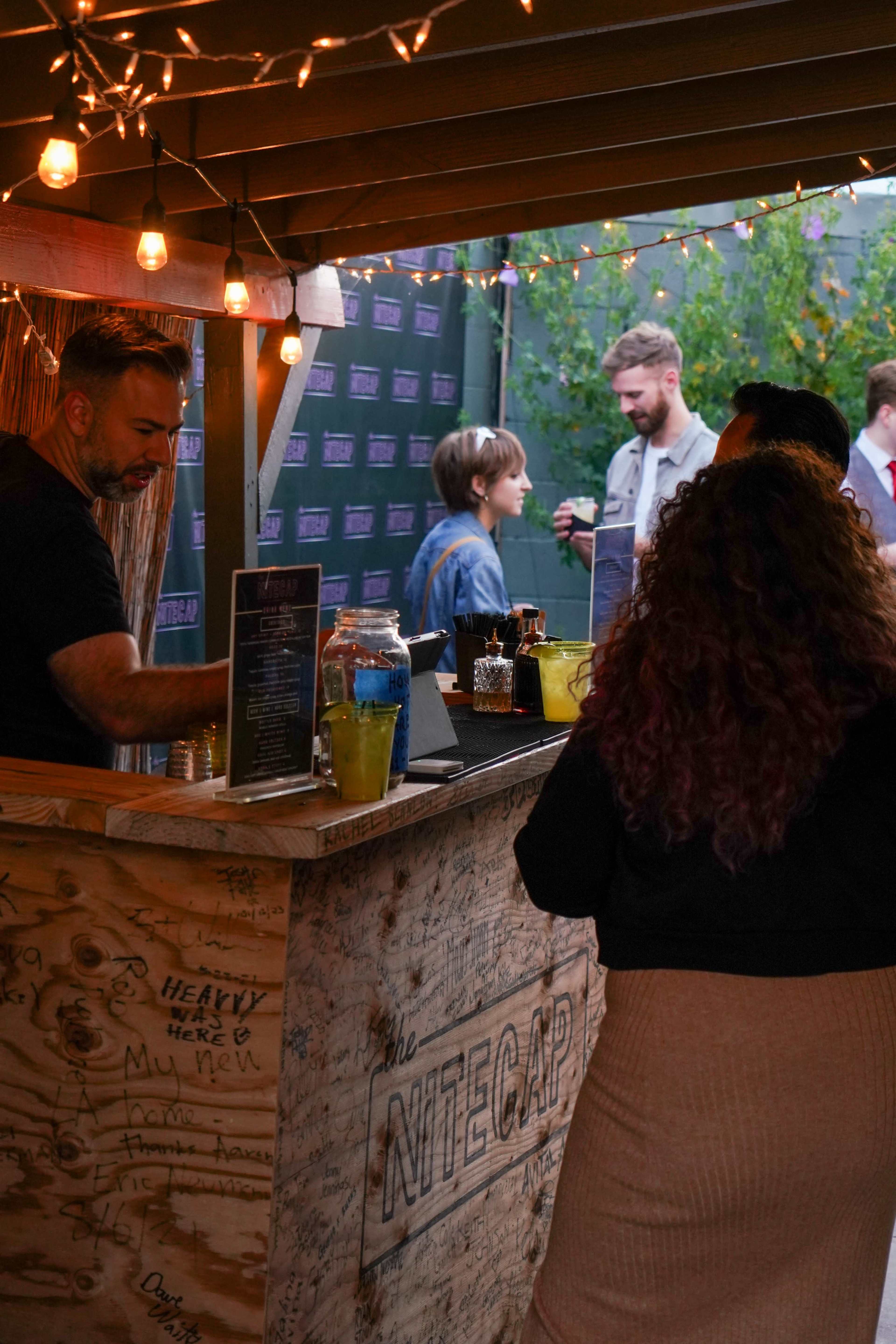 A bartender serves drinks at a rustic bar while patrons gather in an outdoor setting adorned with string lights.
