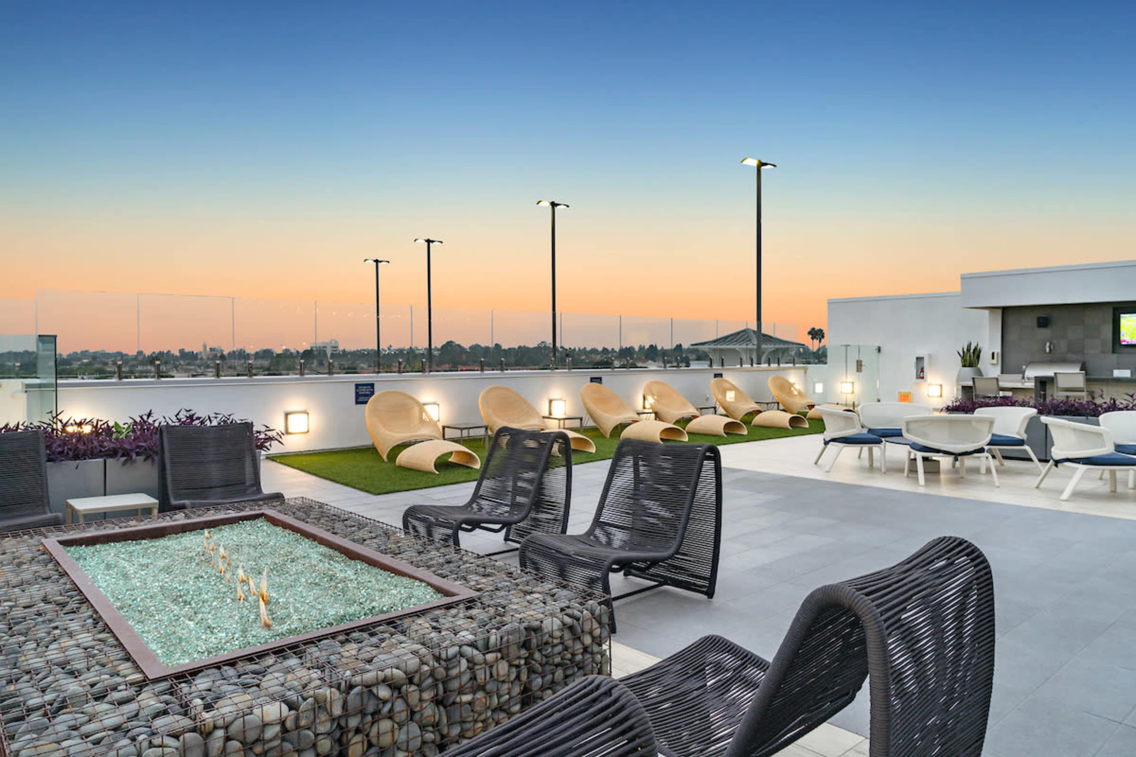 The image shows a modern rooftop lounge area with seating, a fire pit, and a colorful sunset in the background.