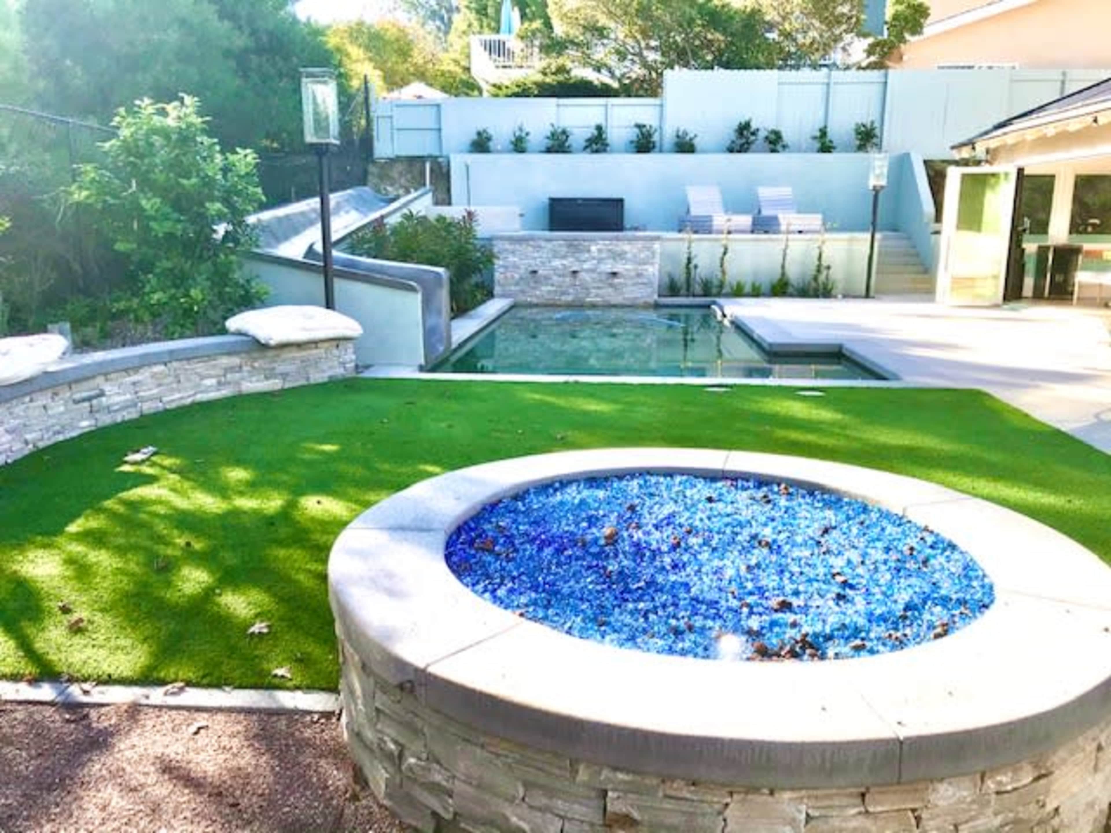 The image shows a landscaped backyard featuring a round fire pit surrounded by blue glass, a green lawn, and a swimming pool with a stone backdrop and lounge chairs.