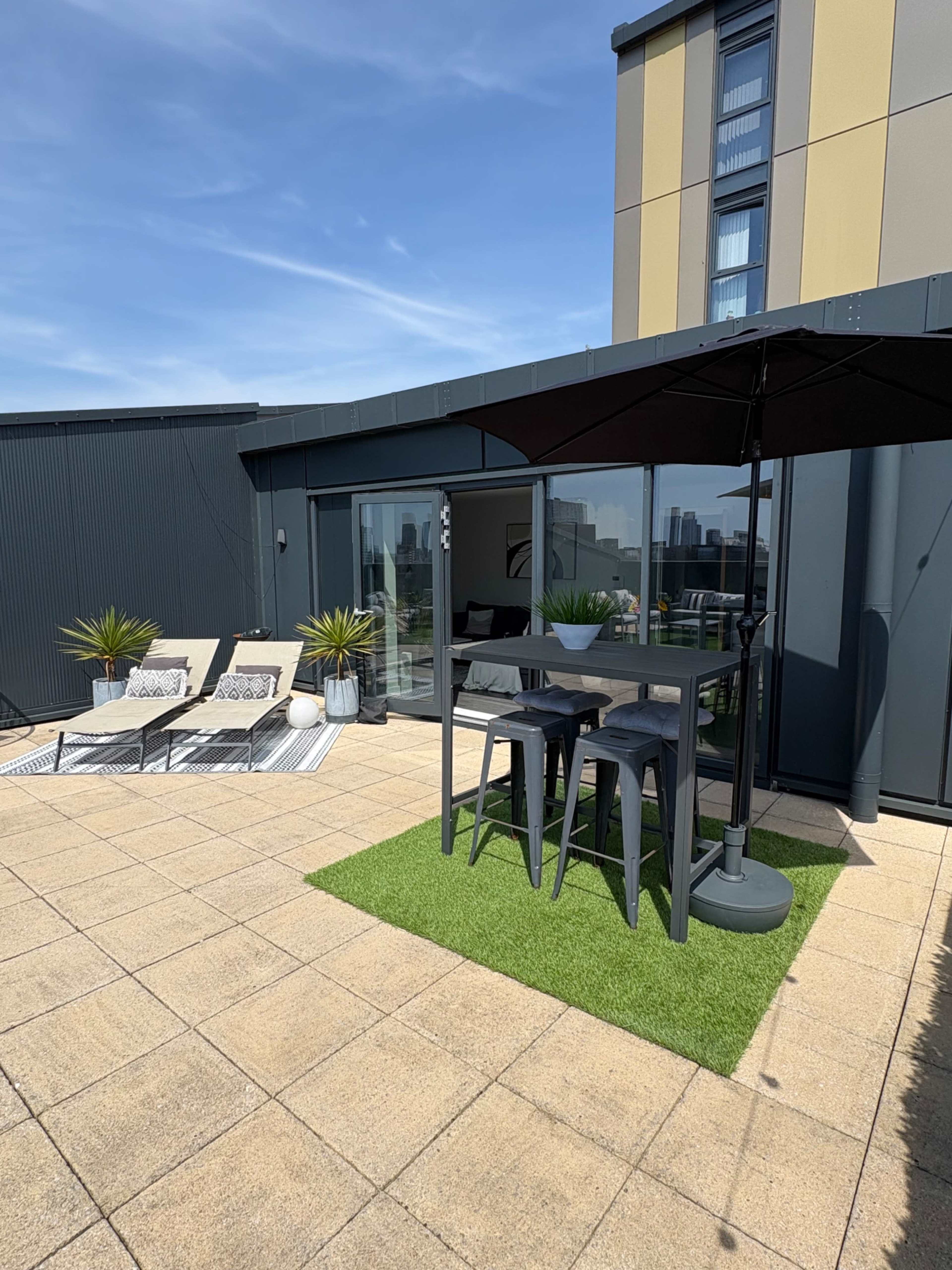 A modern terrace features a black outdoor bar table with stools, surrounded by lounge chairs and potted plants under a clear blue sky.