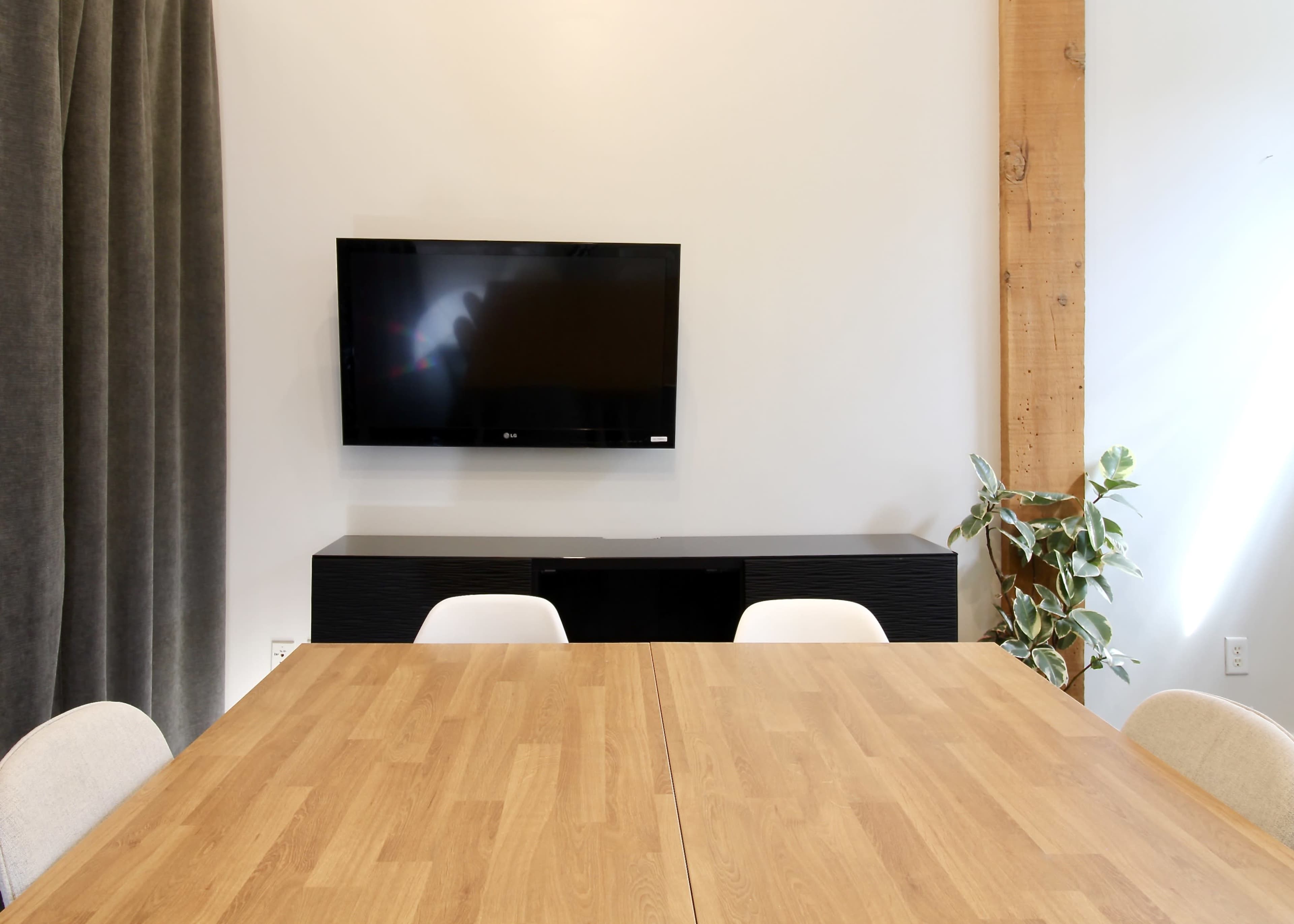 The image shows a modern meeting room with a wooden table, a wall-mounted television, a dark cabinet underneath, and a potted plant beside it.