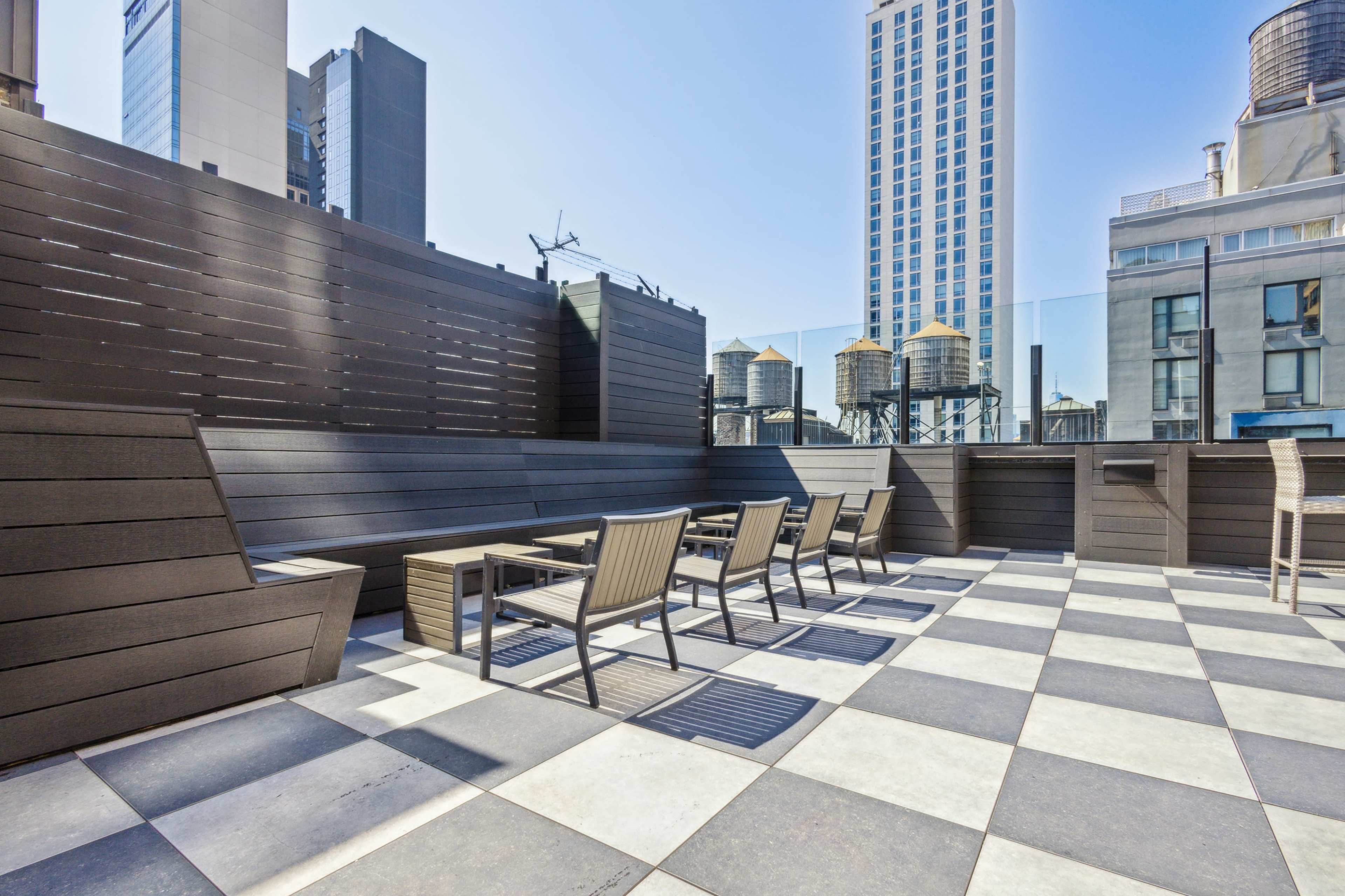 A rooftop terrace with checkered flooring, several lounge chairs, and a view of buildings and water towers in the background under a clear blue sky.