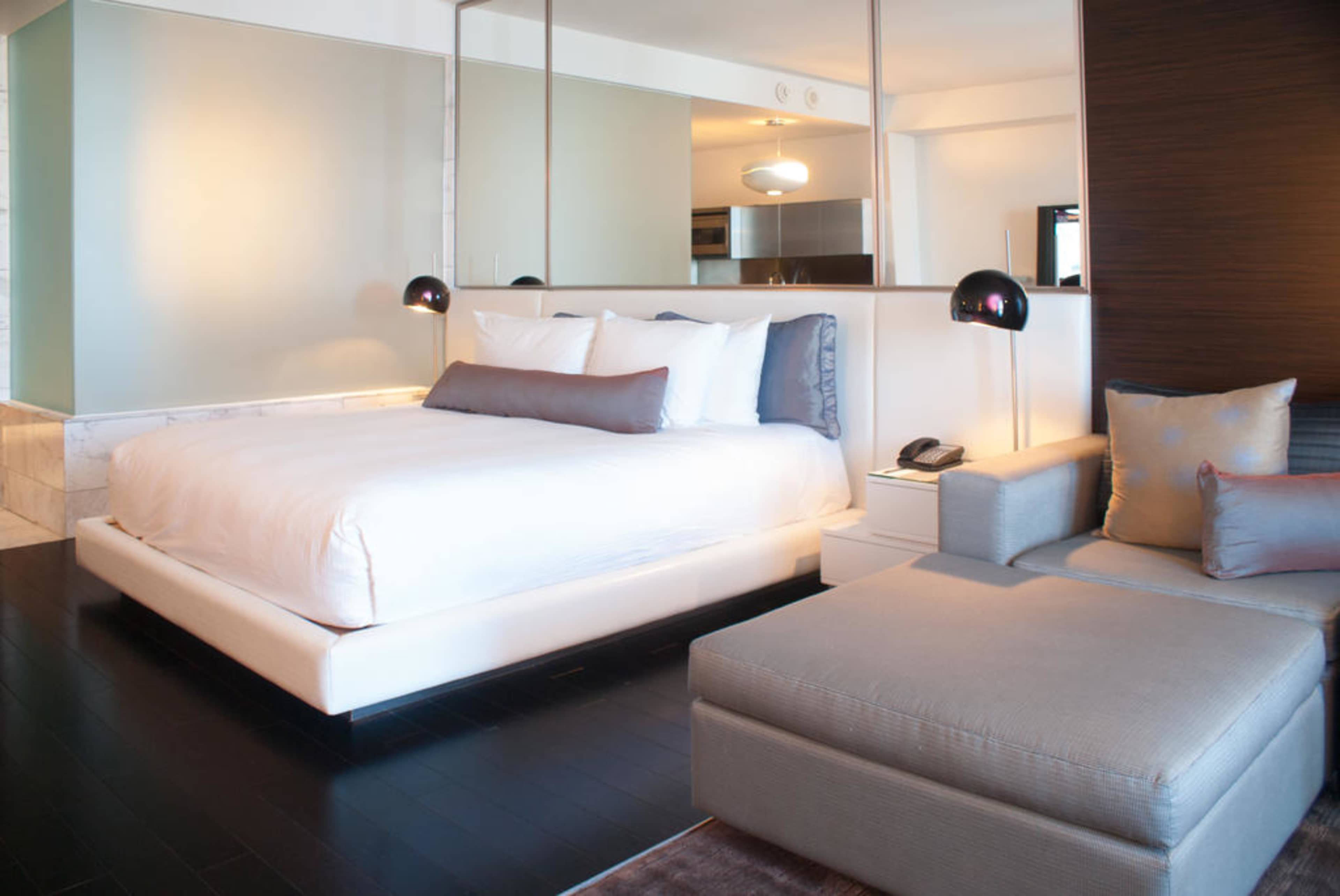 The image shows a modern hotel room featuring a large bed with white linens, a gray chaise lounge, and a mirrored wall.