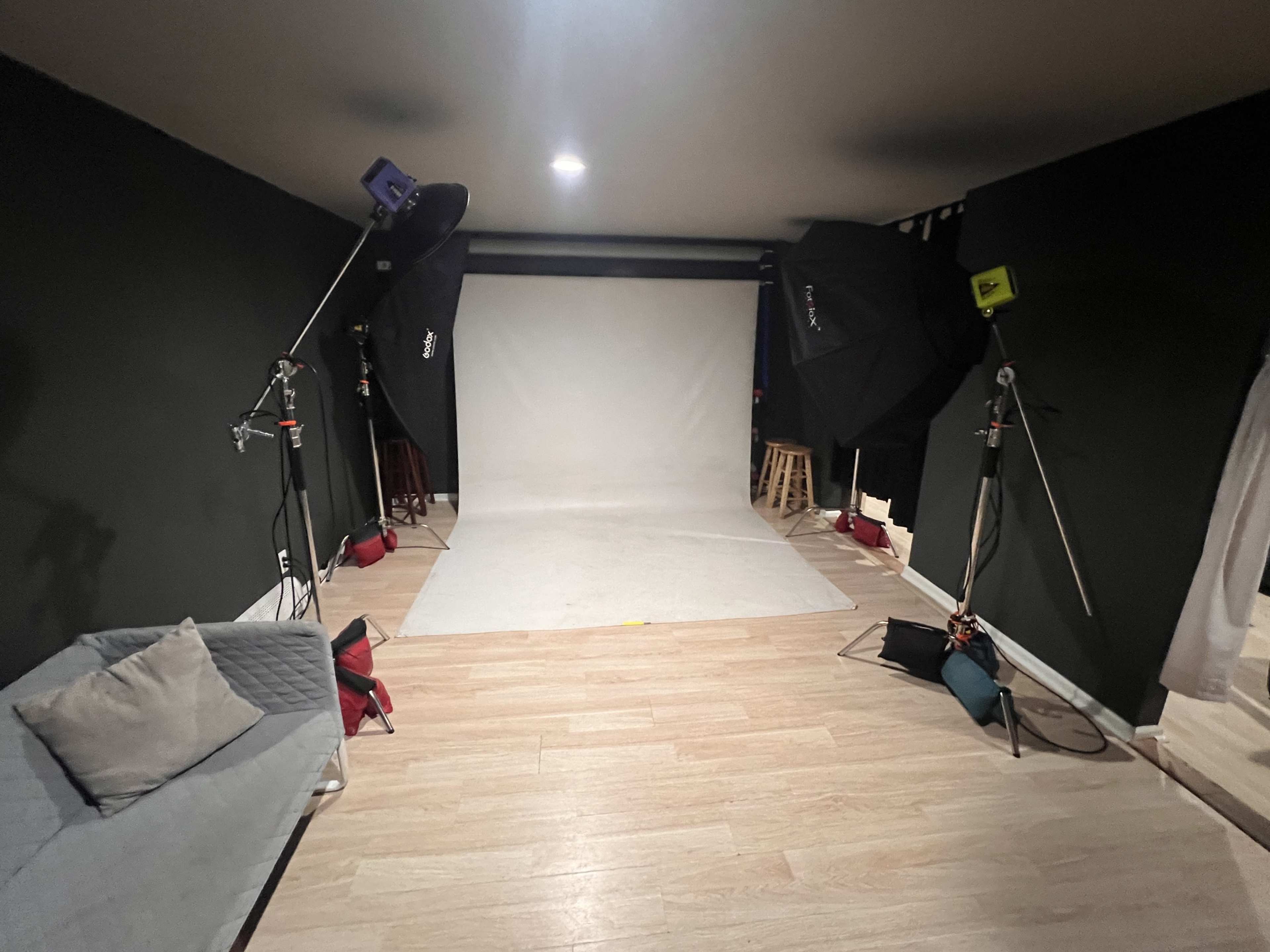 A photography studio with two large softbox lights, a white backdrop, and a gray sofa positioned on a wooden floor.