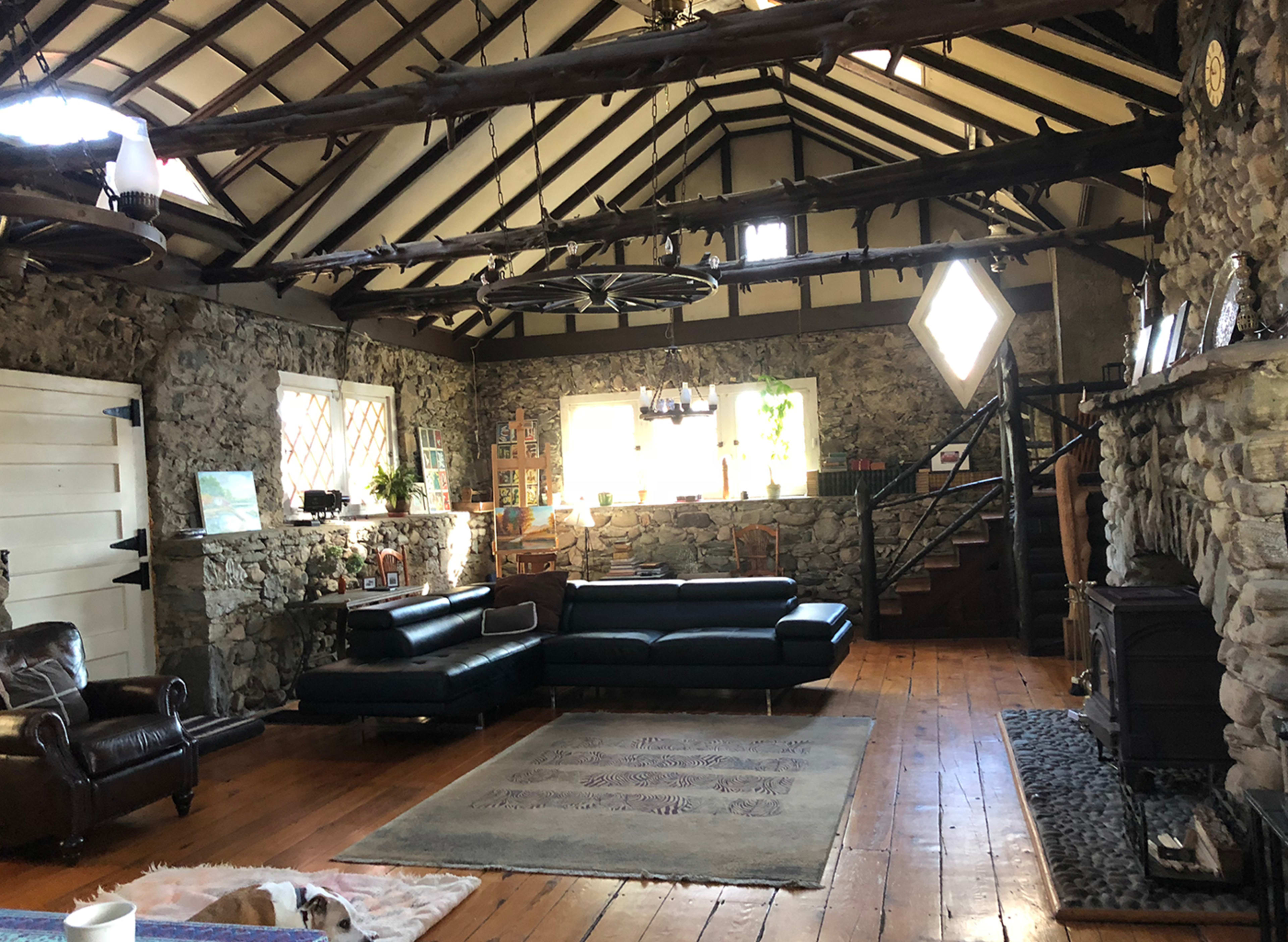 The image shows a rustic living room with stone walls, wooden beams, and a mix of seating options, including a black leather sectional and a brown armchair.
