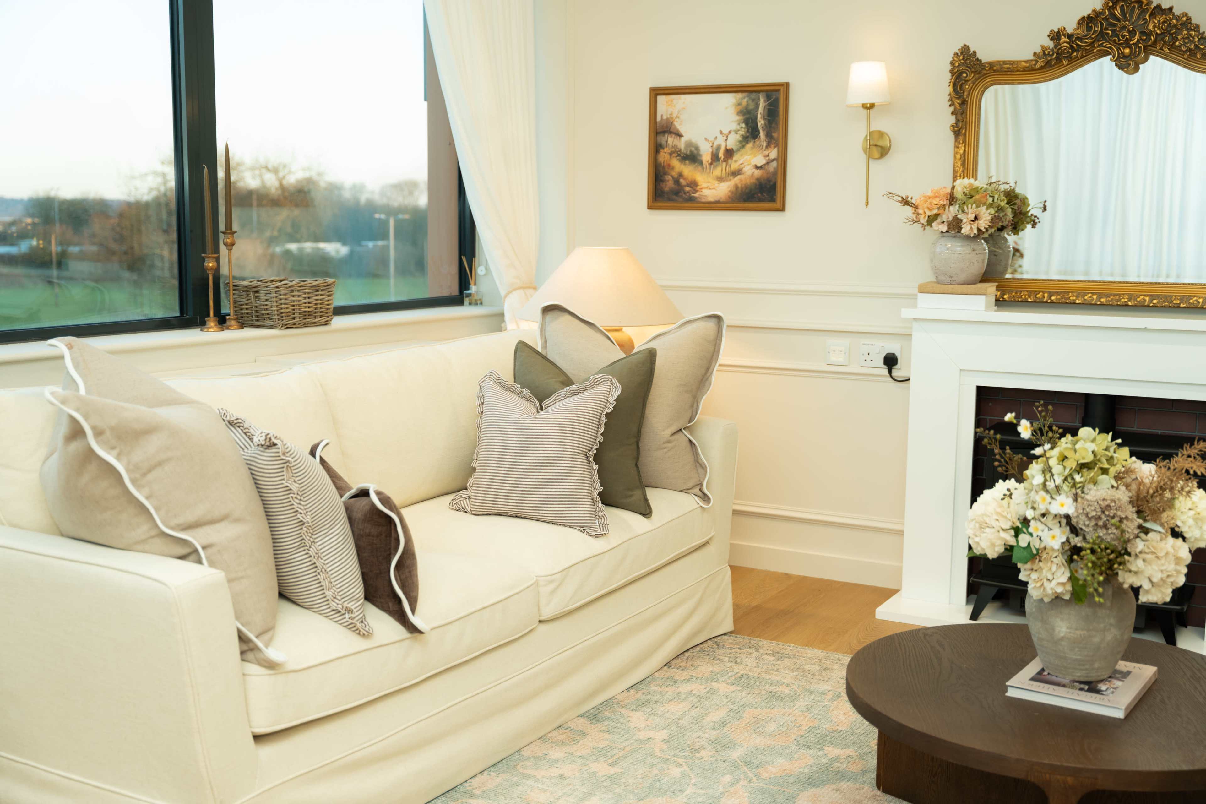 The image shows a cozy living room scene featuring a light-colored sofa adorned with various throw pillows, a small table with a floral arrangement, and a mirror above a decorative fireplace.