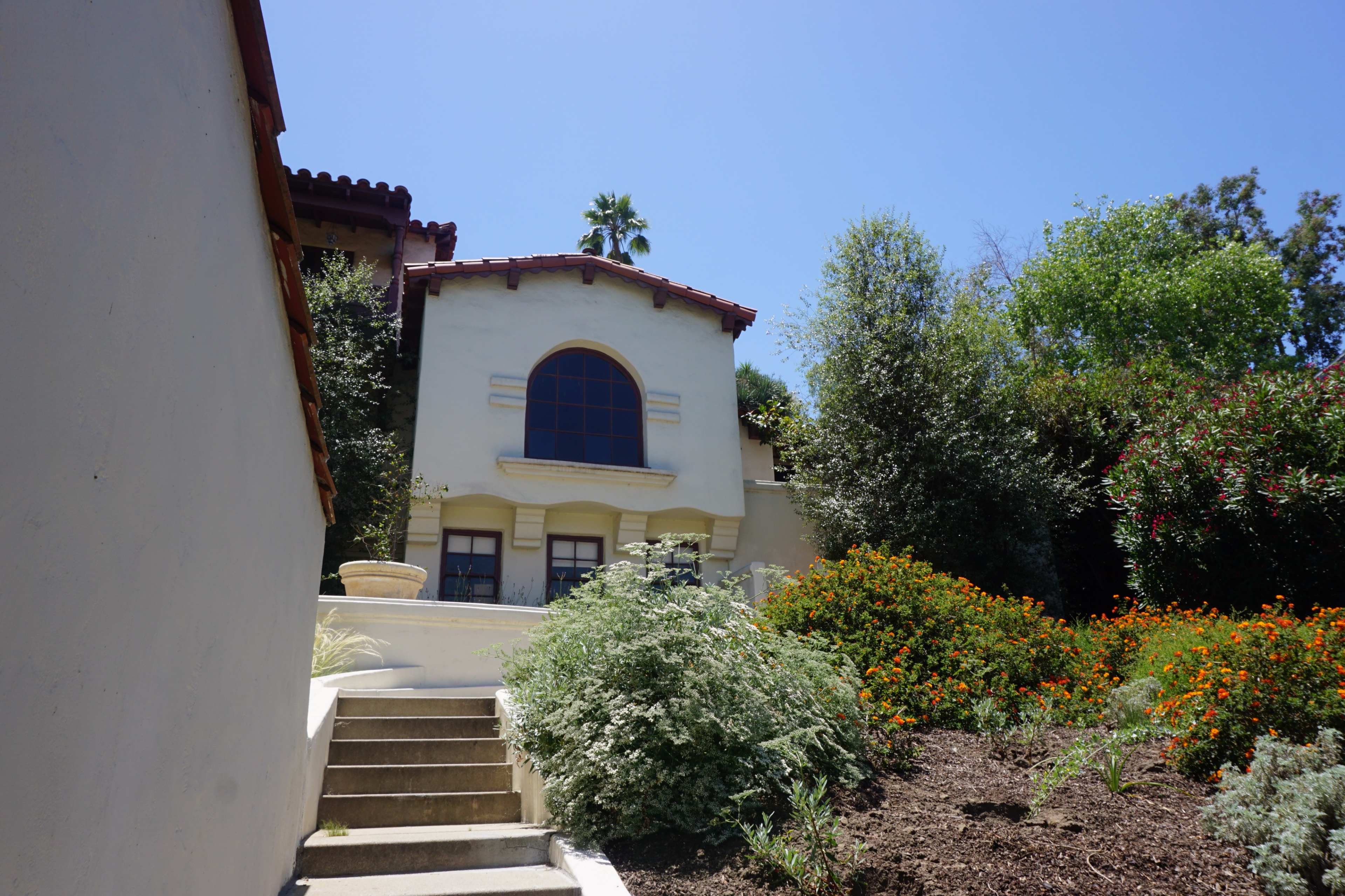 A two-story house with a red-tiled roof is surrounded by greenery and colorful flowers, with a set of stairs leading up to its entrance.