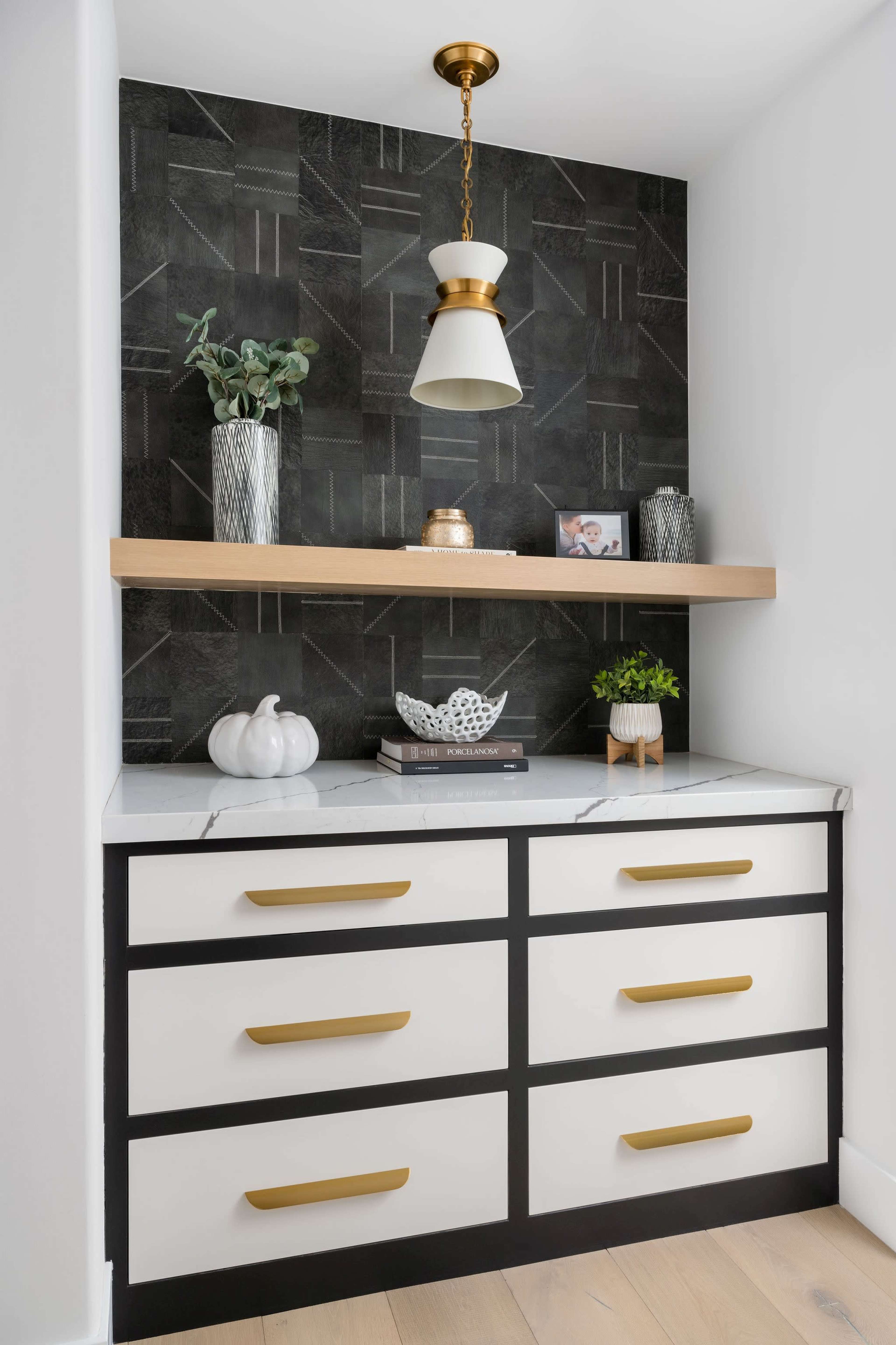 A modern cabinetry setup features a dark wall with geometric patterns, a wooden shelf, and decorative items, including a white pumpkin and plants, above a white and black dresser with gold handles.