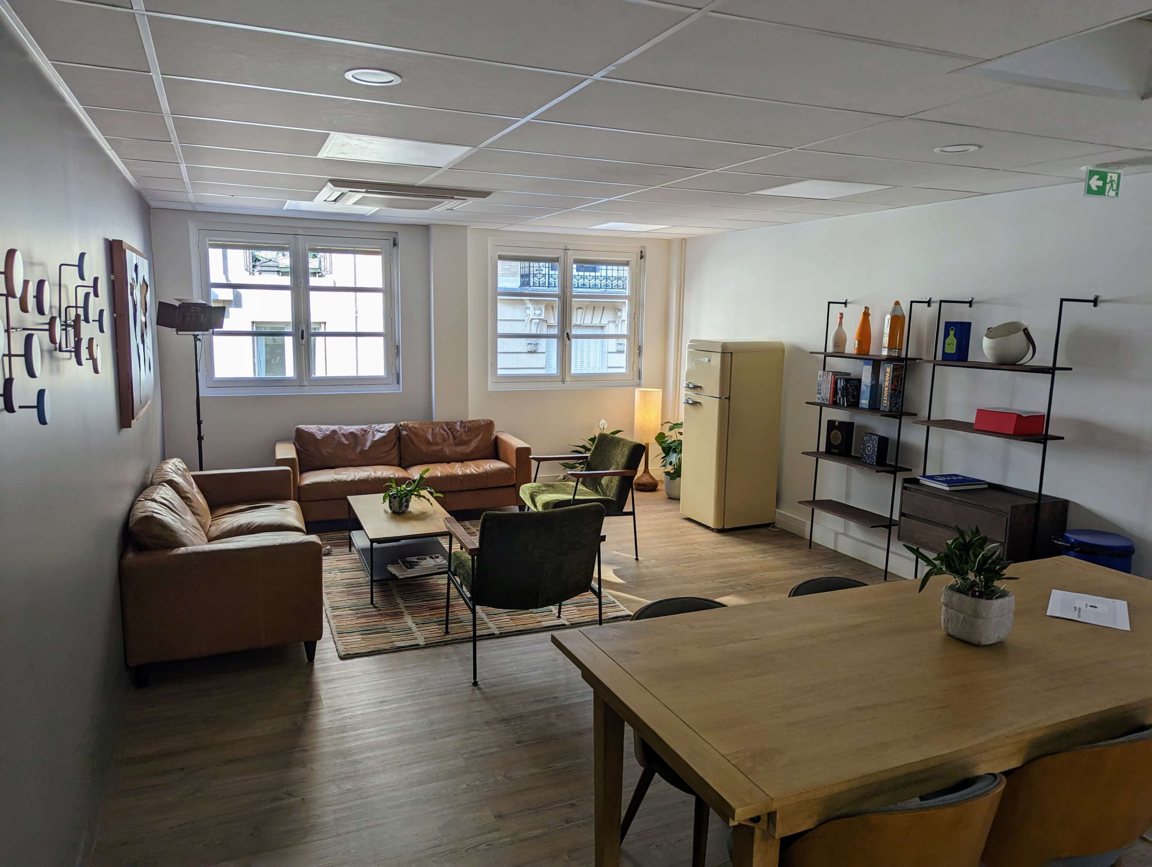 A modern office lounge area with a brown leather sofa, a coffee table, a dining table, and a vintage-style refrigerator against a light-colored wall.