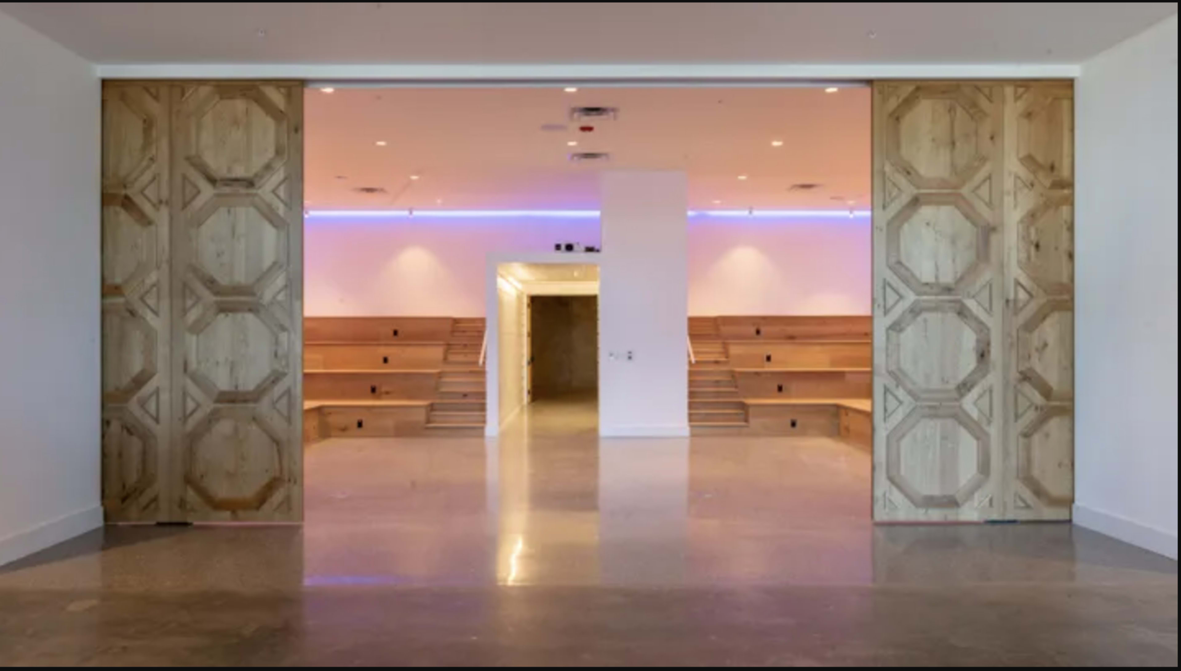 The image shows a spacious interior with large wooden double doors leading to an area featuring tiered wooden seating and illuminated walls.