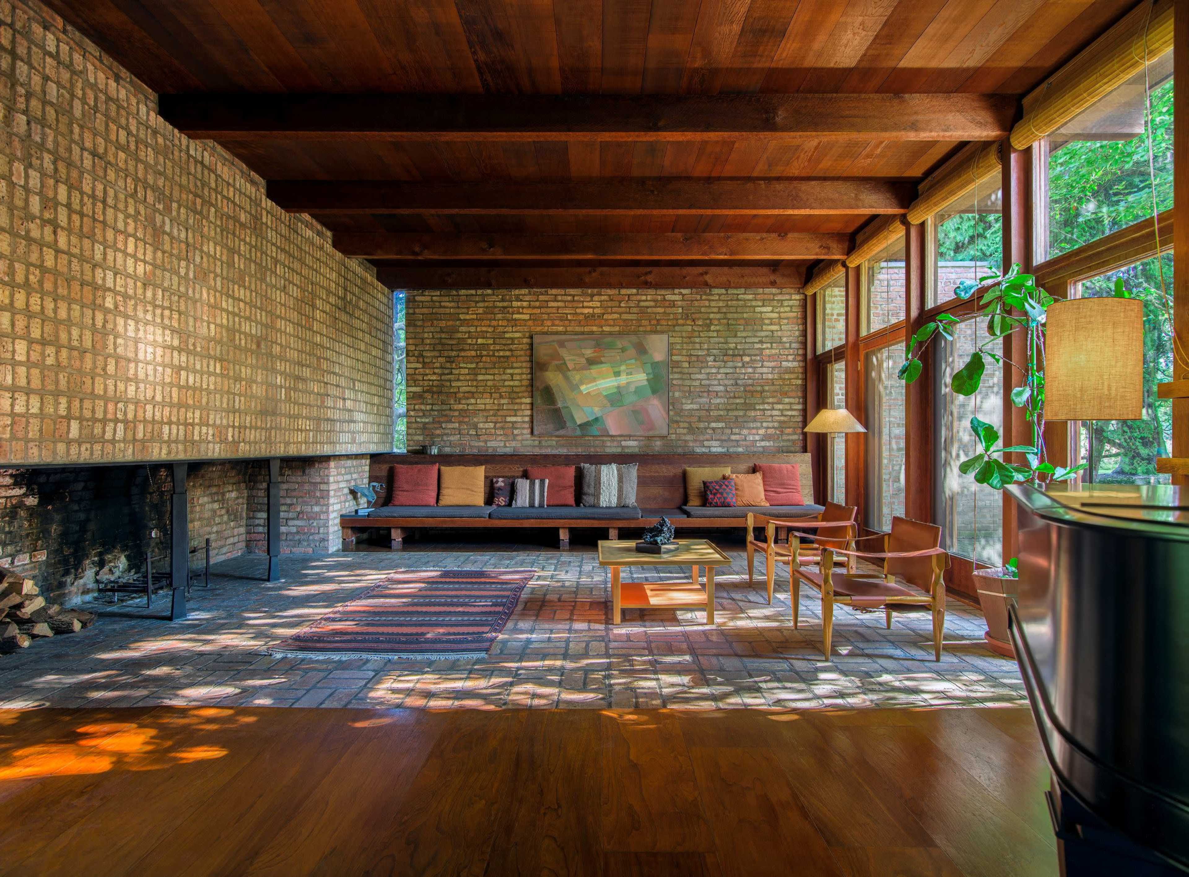 A spacious living room features a brick wall, wooden beams, large windows, and minimal furniture, including a sofa, chairs, and a lamp.