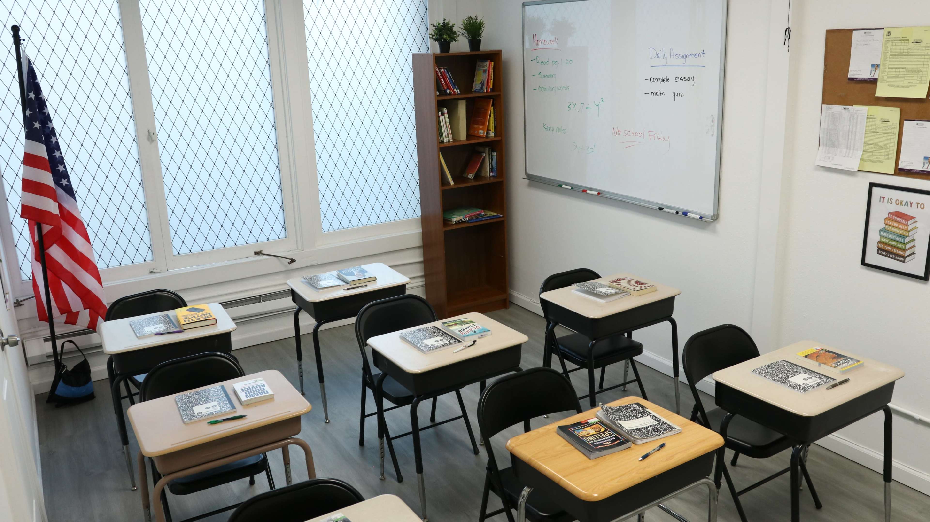 The image shows a classroom with several desks arranged in rows, an American flag in the corner, a bookshelf filled with books, and a whiteboard displaying notes.