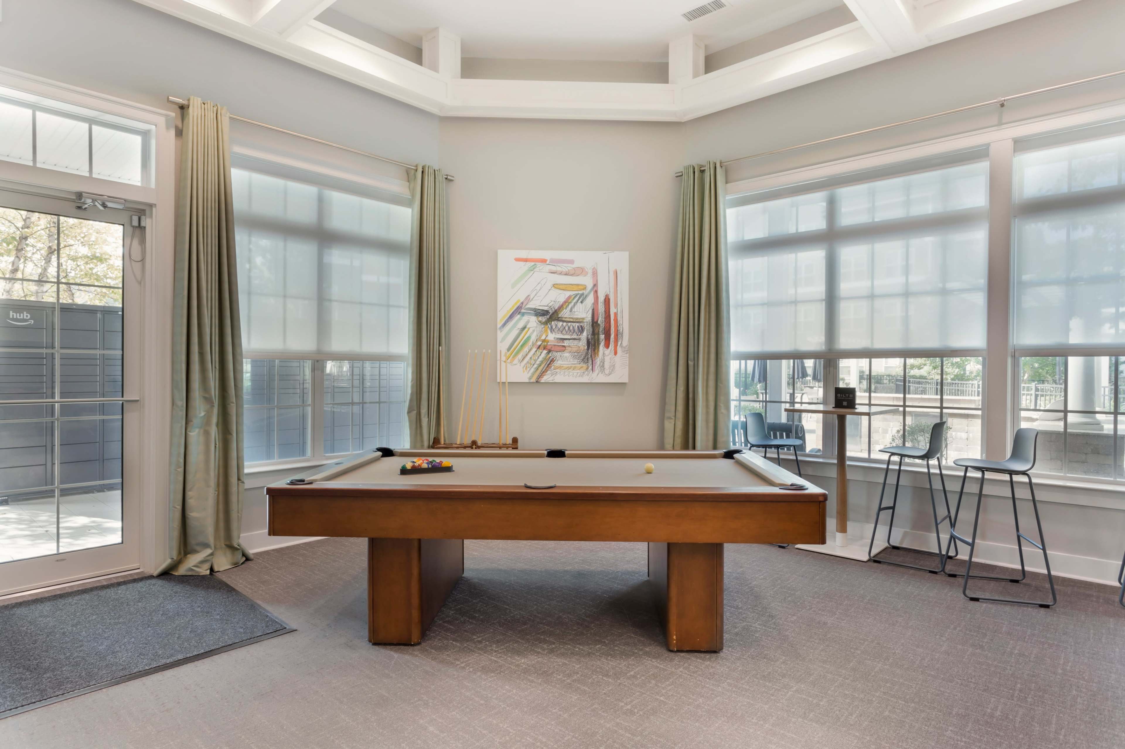 The image shows a well-lit room featuring a pool table, large windows with sheer curtains, and a small dining set in the corner.