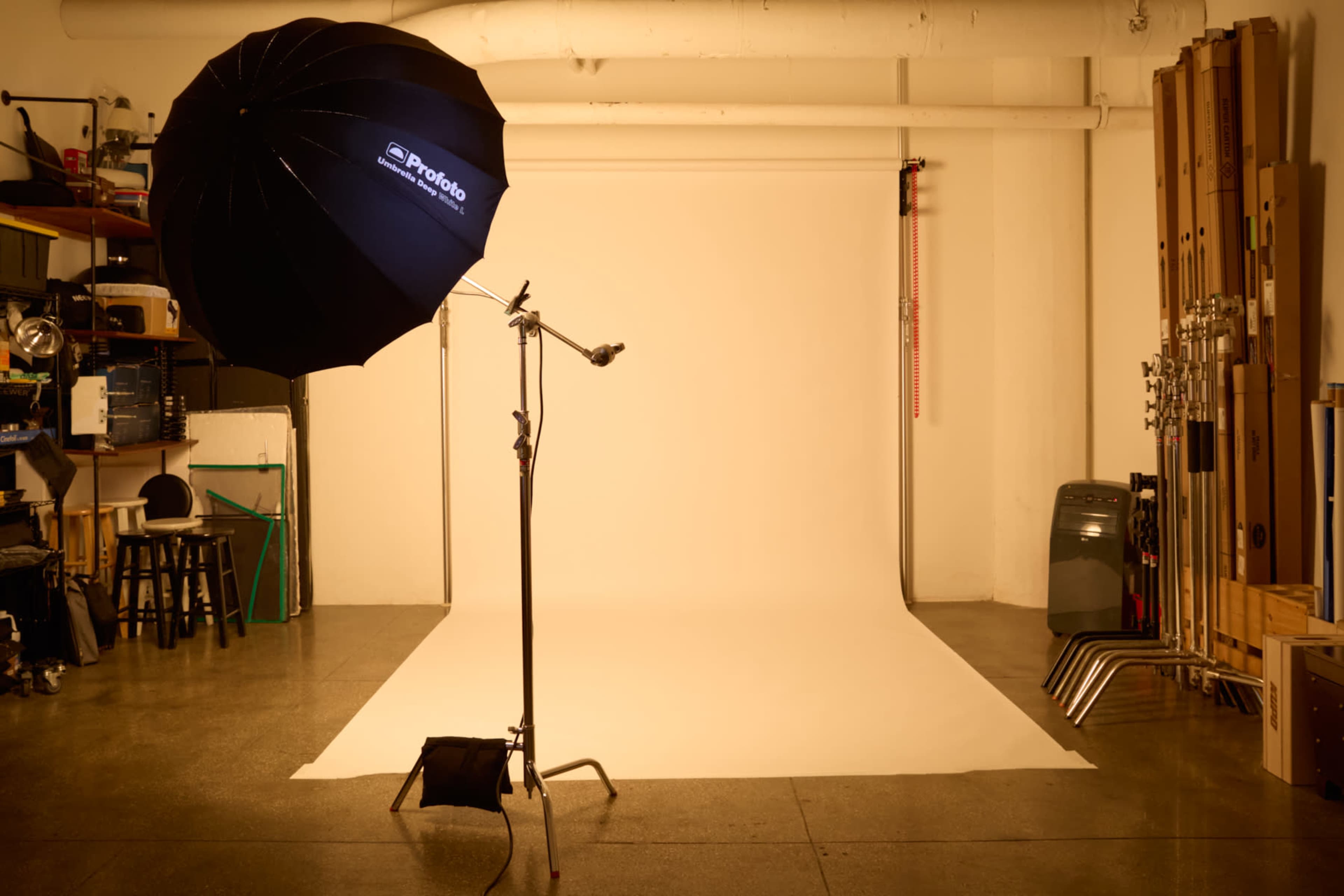 A photography studio features a softbox umbrella light on a stand, positioned in front of a blank backdrop with various equipment stored along the walls.