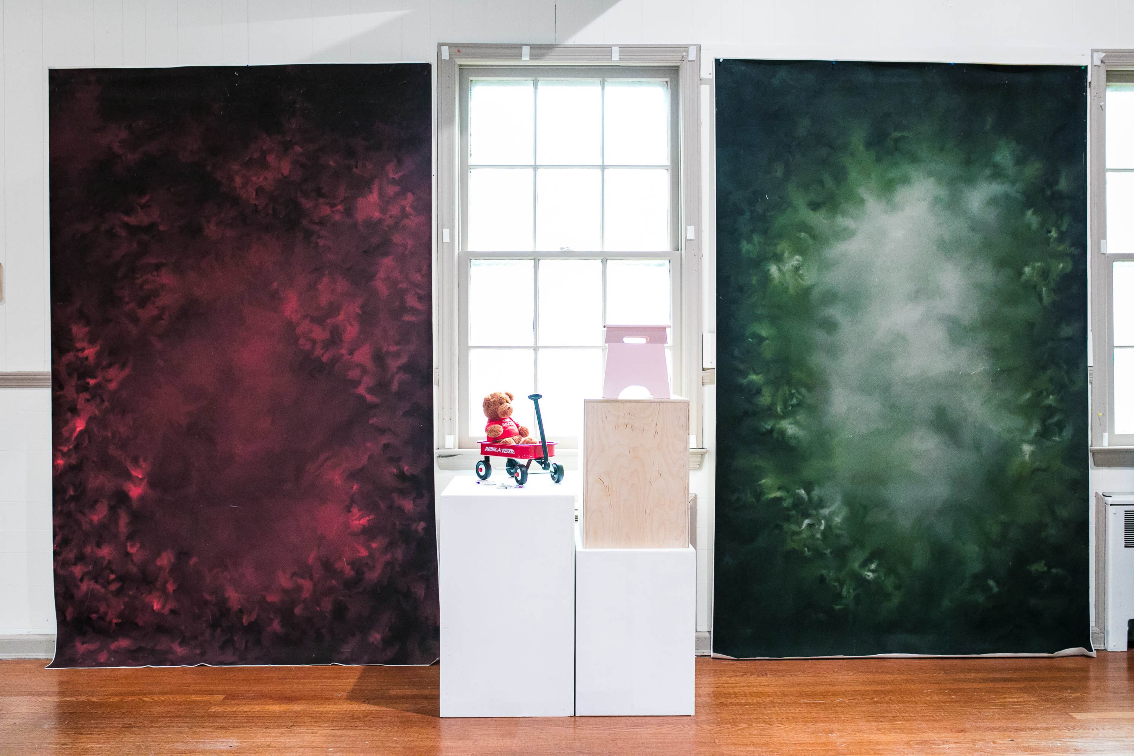 Two large, colorful fabric hangings on walls flanking a central white display pedestal with a toy bear sitting on a red toy wagon.