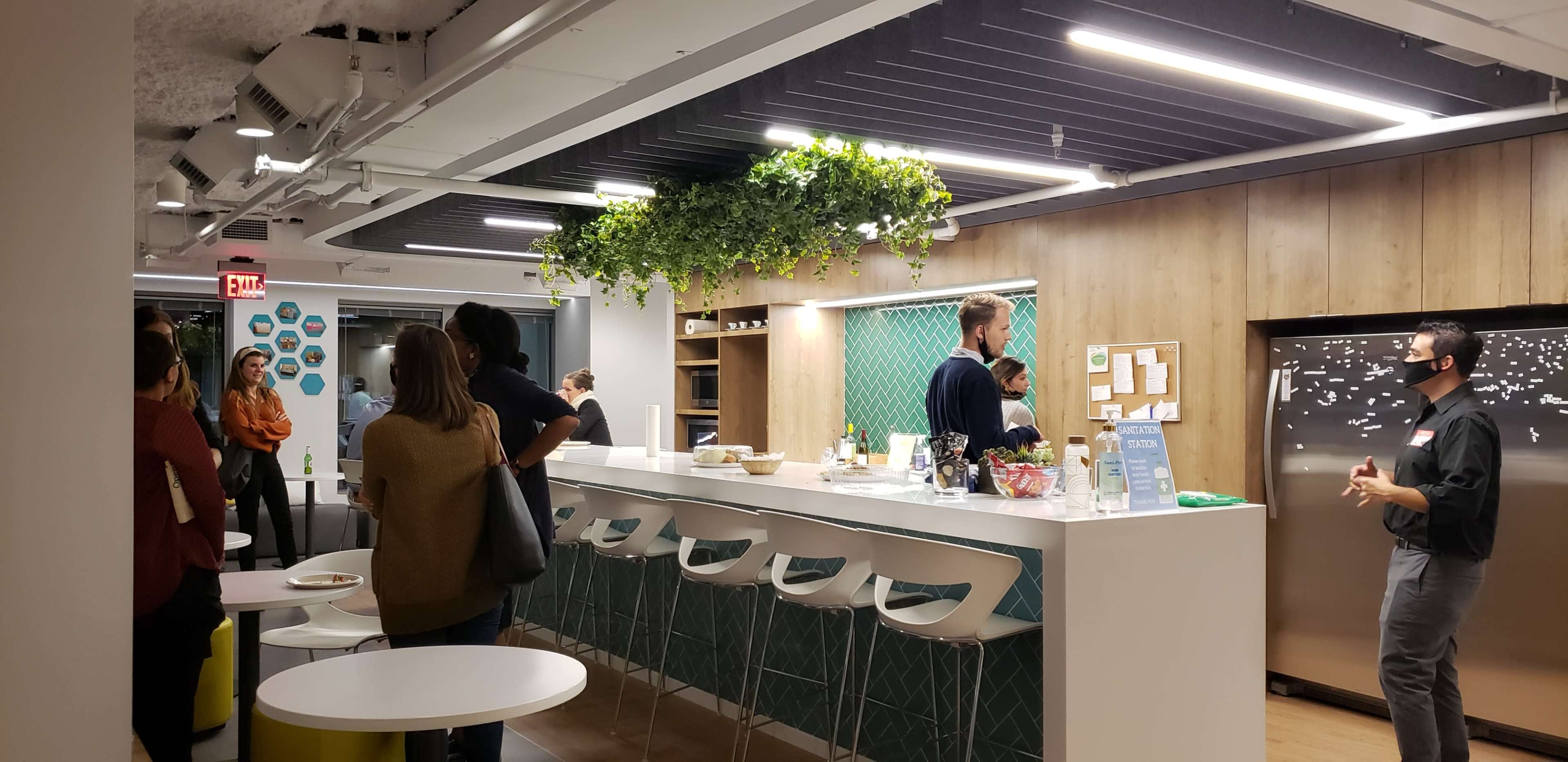A group of people engage in conversation in a modern kitchen area featuring a large counter, greenery, and a refrigerator.