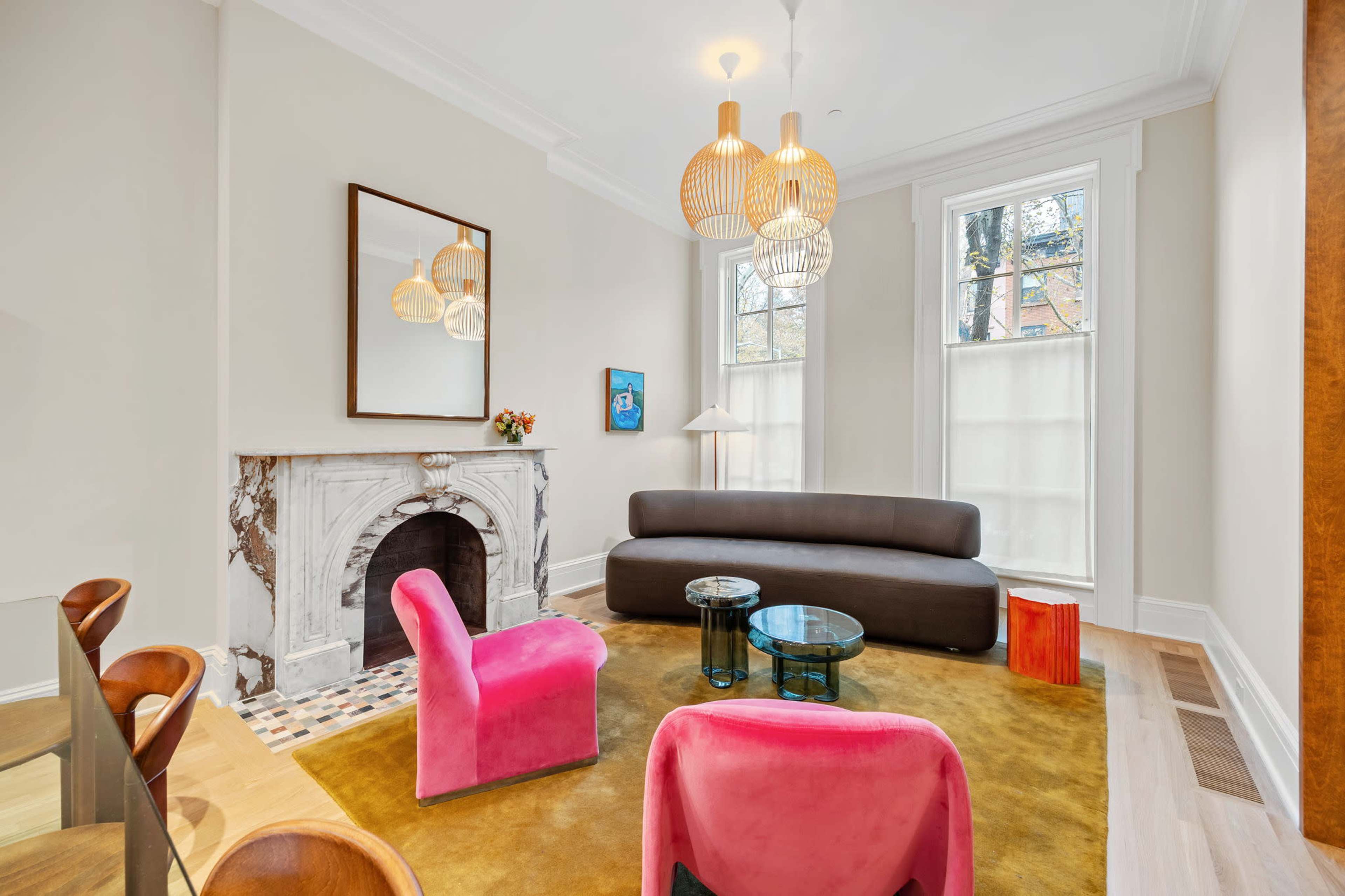 The image shows a modern living room featuring a large brown sofa, two pink chairs, a round glass coffee table, and a marble fireplace, with large windows letting in natural light.