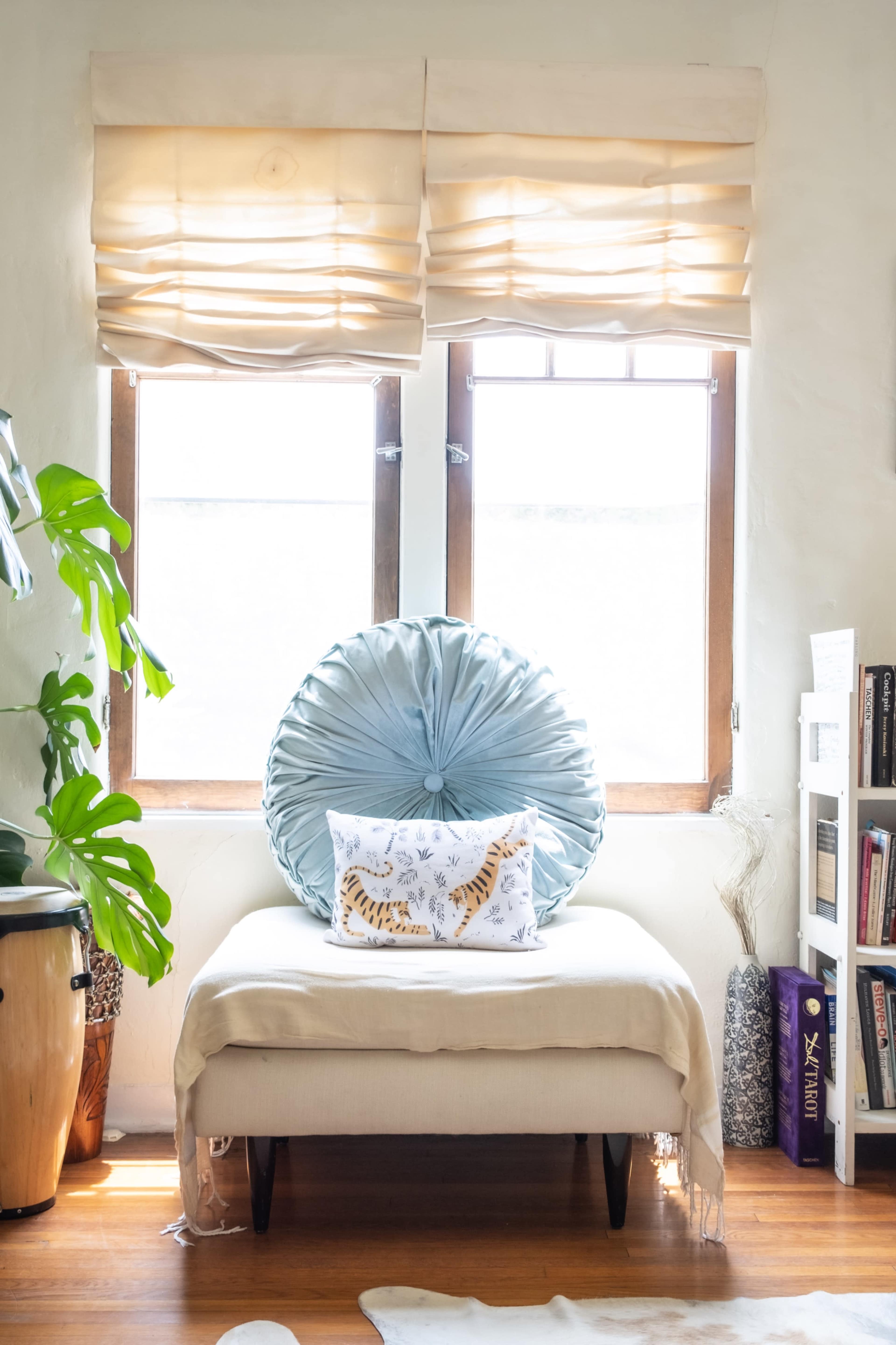 A light-filled room features a tufted blue cushion and a decorative pillow on a bench beneath two windows with fabric shades, alongside a potted plant and a bookshelf.