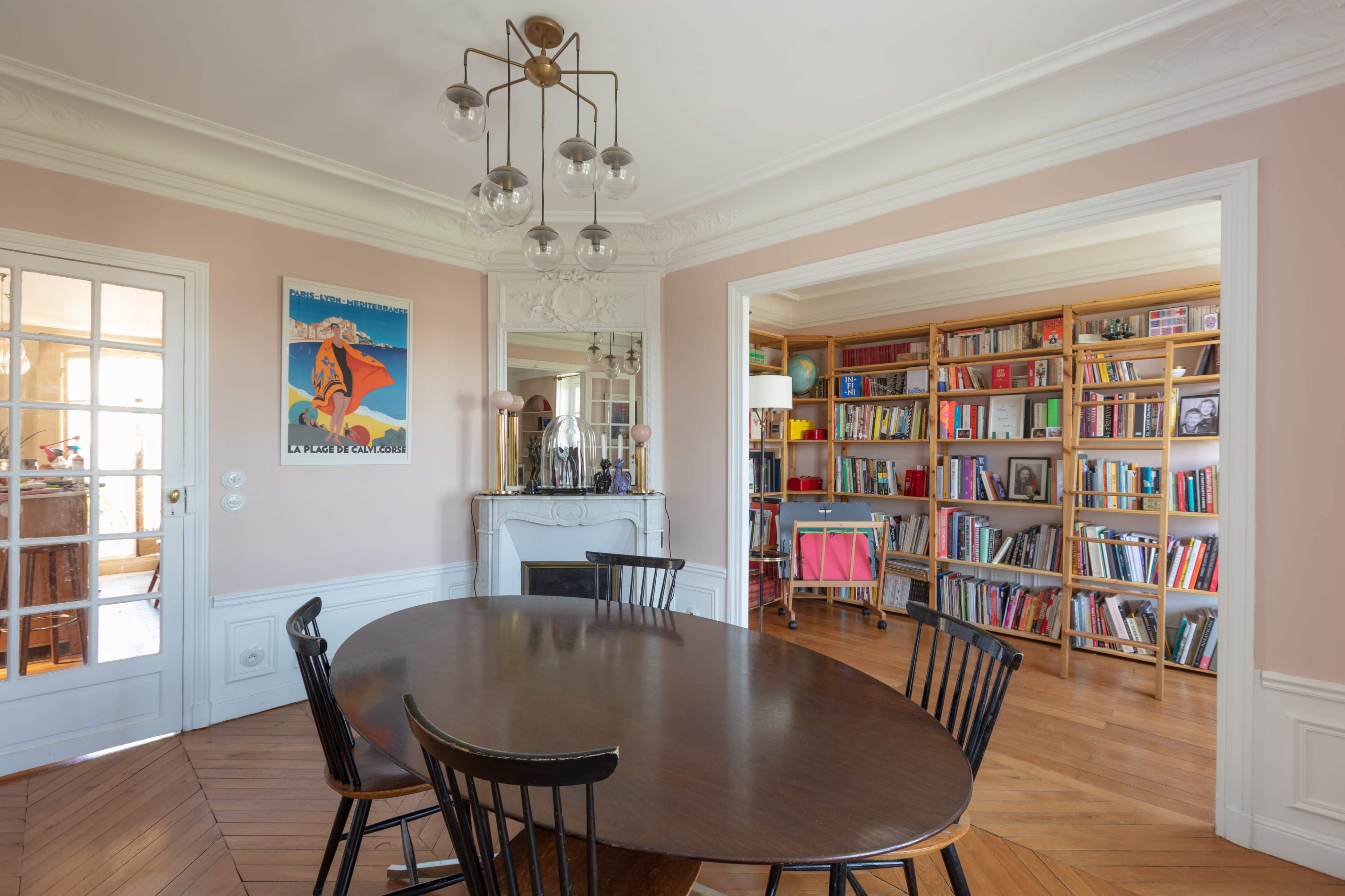 A round wooden dining table with black chairs is set in a light-colored room that connects to a library filled with bookshelves and an ornate fireplace.