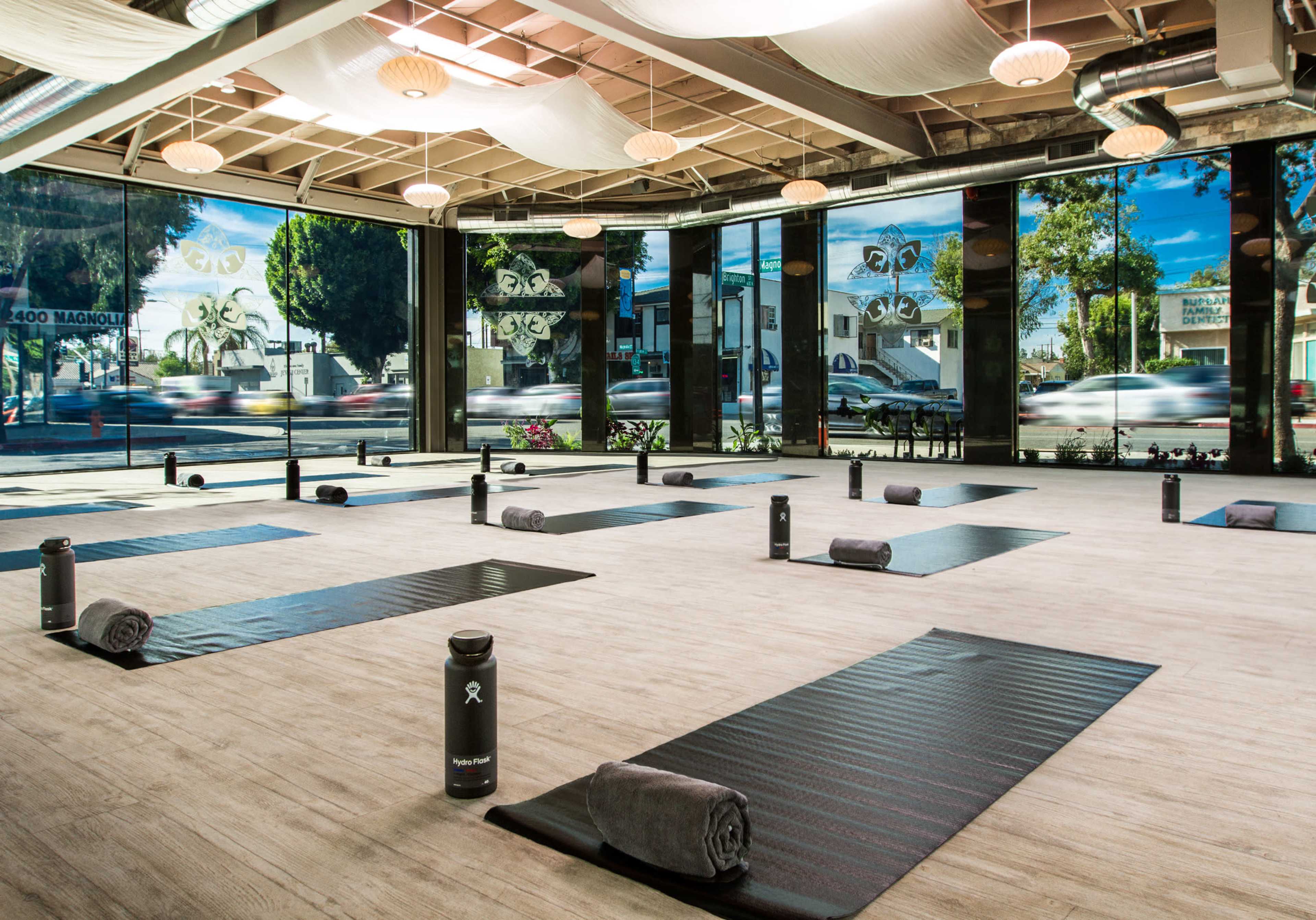 The image shows a fitness studio with neatly arranged yoga mats and water bottles, illuminated by natural light through large windows.