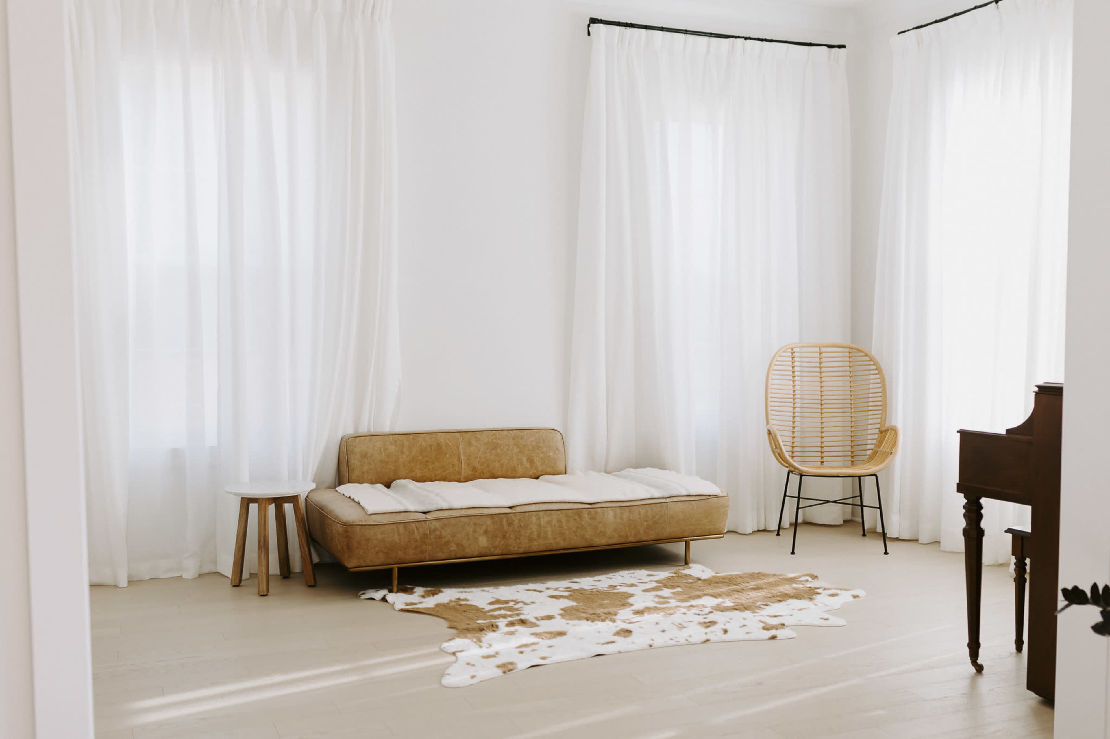 A minimalist living room featuring a brown sofa, a small wooden side table, a rattan chair, a piano, and light curtains.