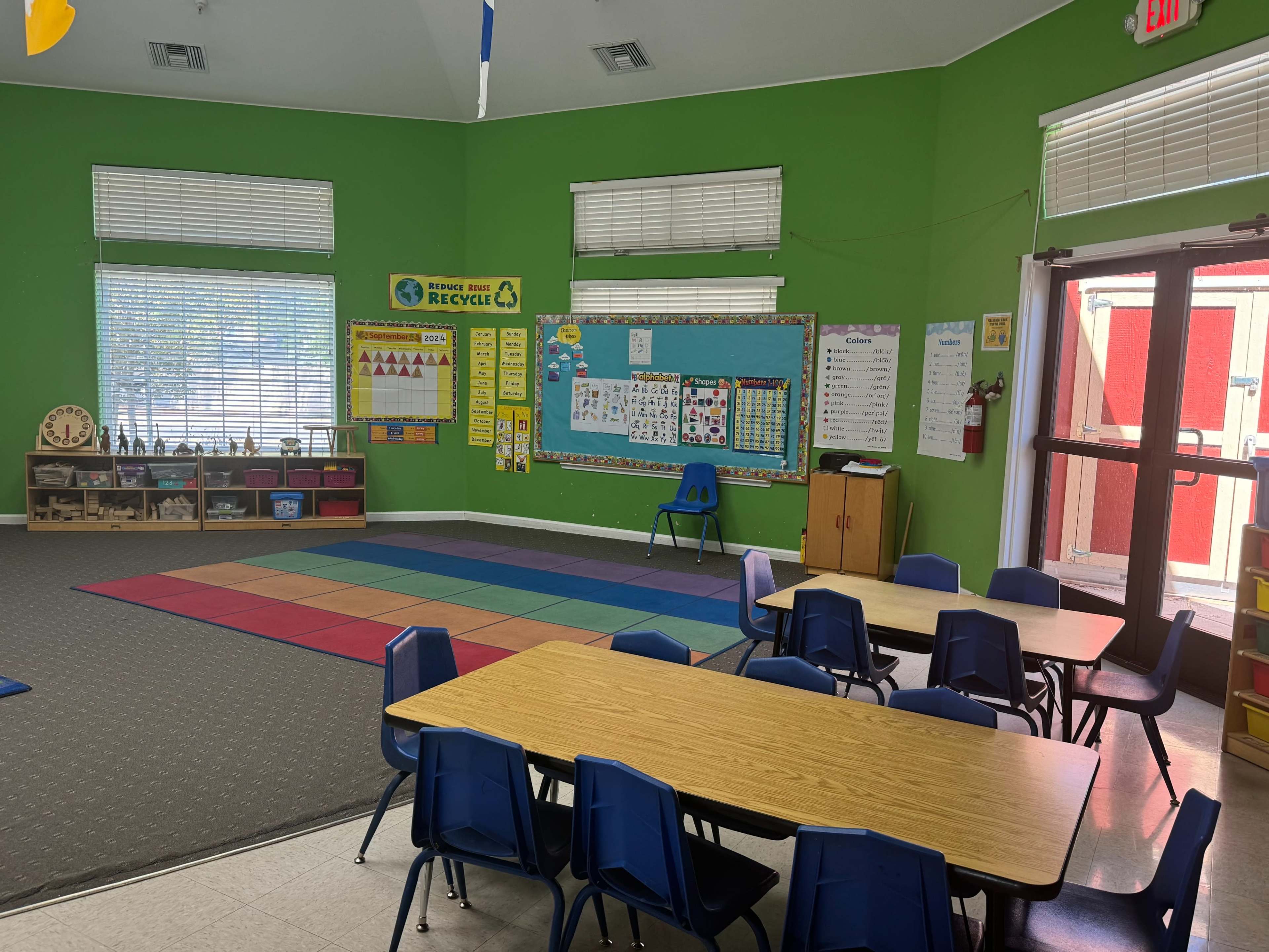 The classroom features a green wall, colorful floor mats, tables with chairs, and educational materials displayed on bulletin boards.